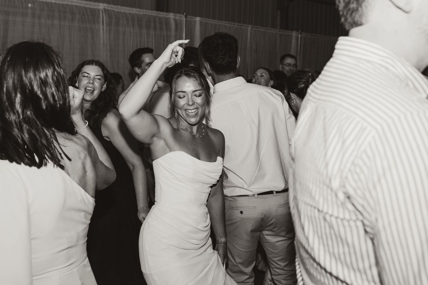 Black and white photo of a bride in a white strapless dress dancing joyfully with guests at a wedding reception.