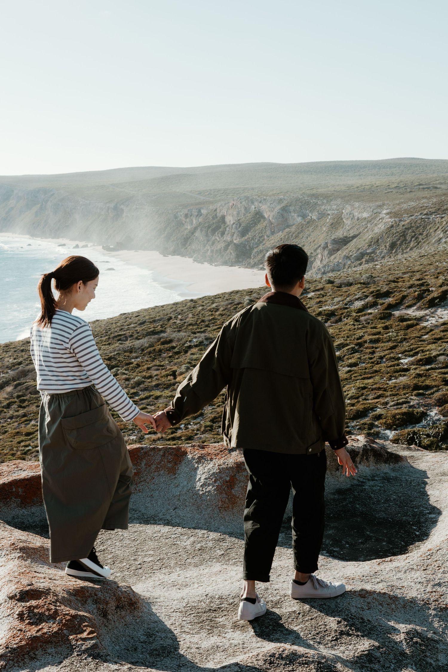 Couple holding hands while standing on coastal cliff overlooking dramatic ocean views.