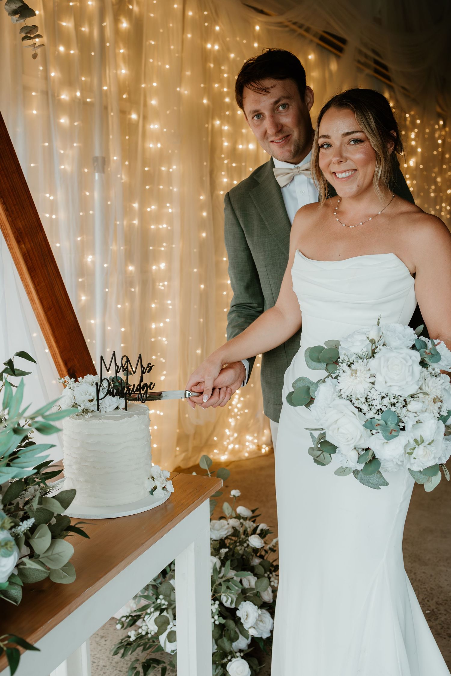 Bride and groom cutting white wedding cake with fairy lights backdrop and white floral bouquet.