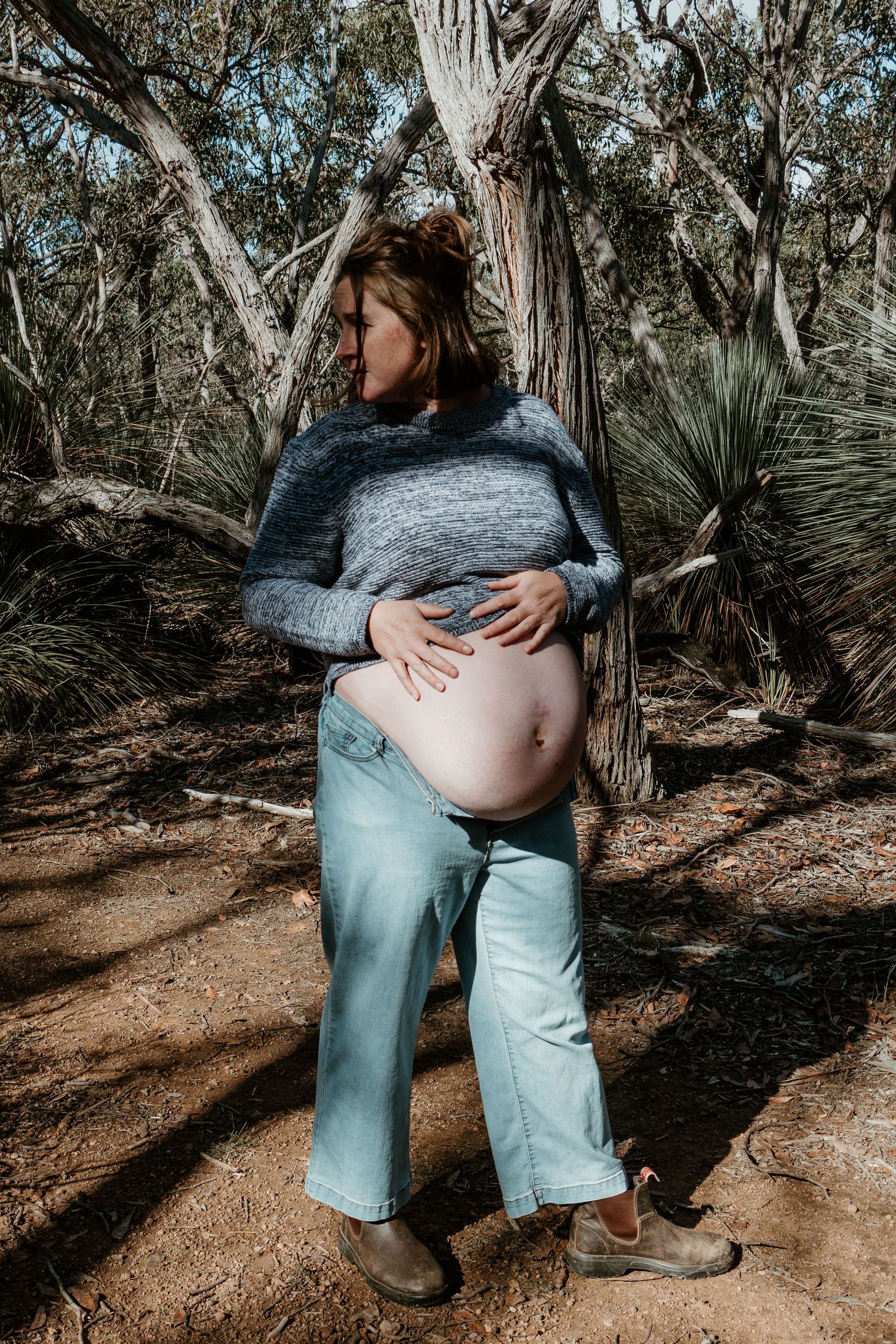 Pregnant woman in grey sweater and jeans cradling baby bump outdoors among eucalyptus trees in natural bush setting.