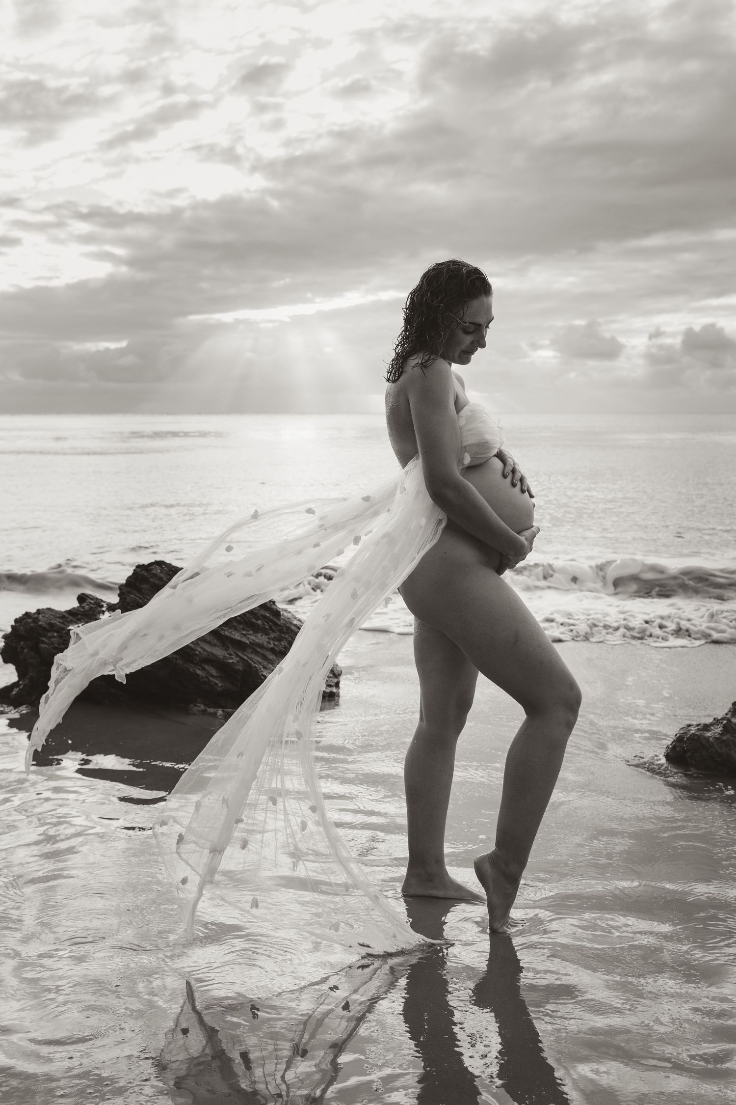 A silhouetted pregnant figure draped in flowing fabric stands on a beach at sunset with ocean waves and cloudy sky.
