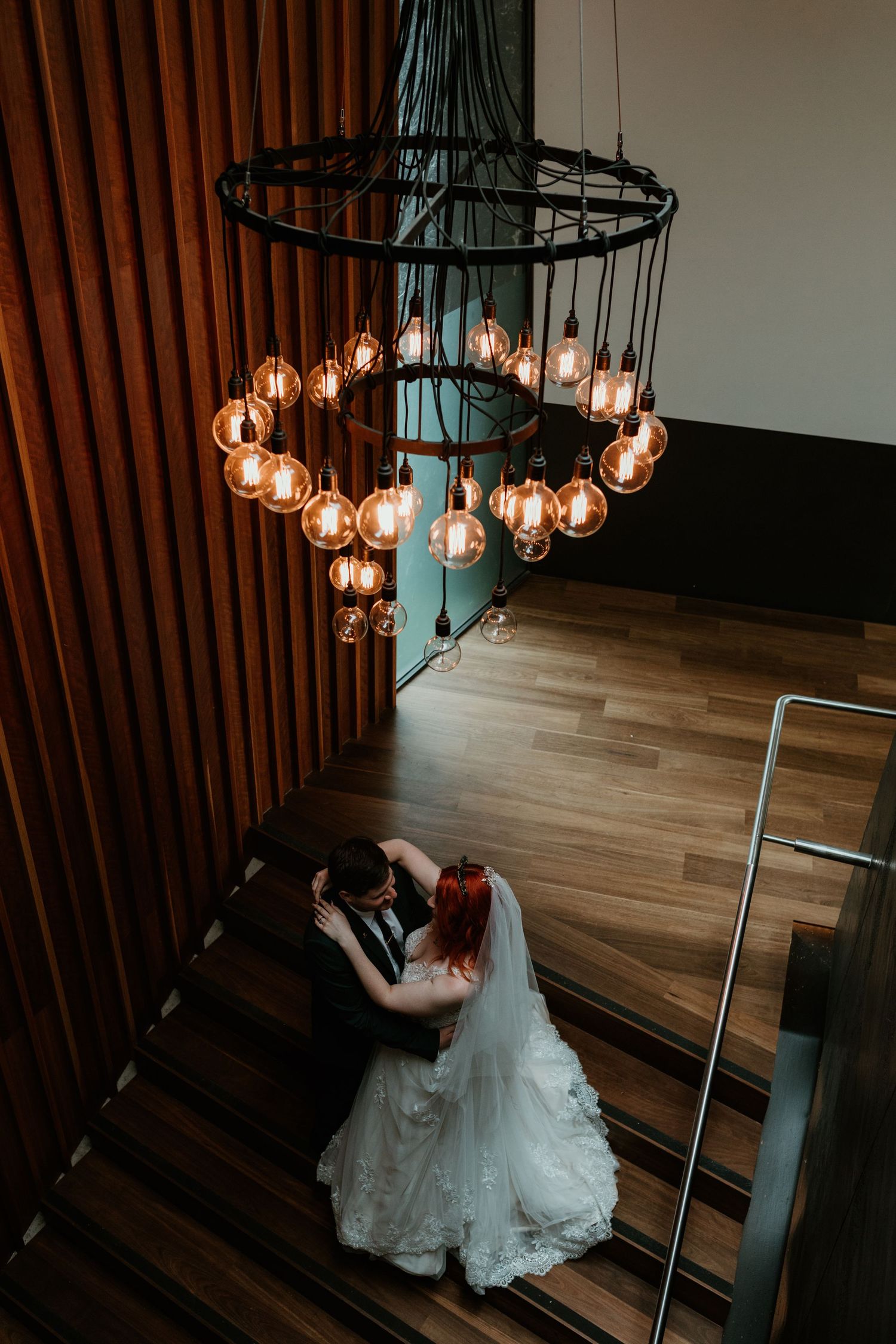 A wedding couple embracing on stairs beneath a striking circular chandelier.