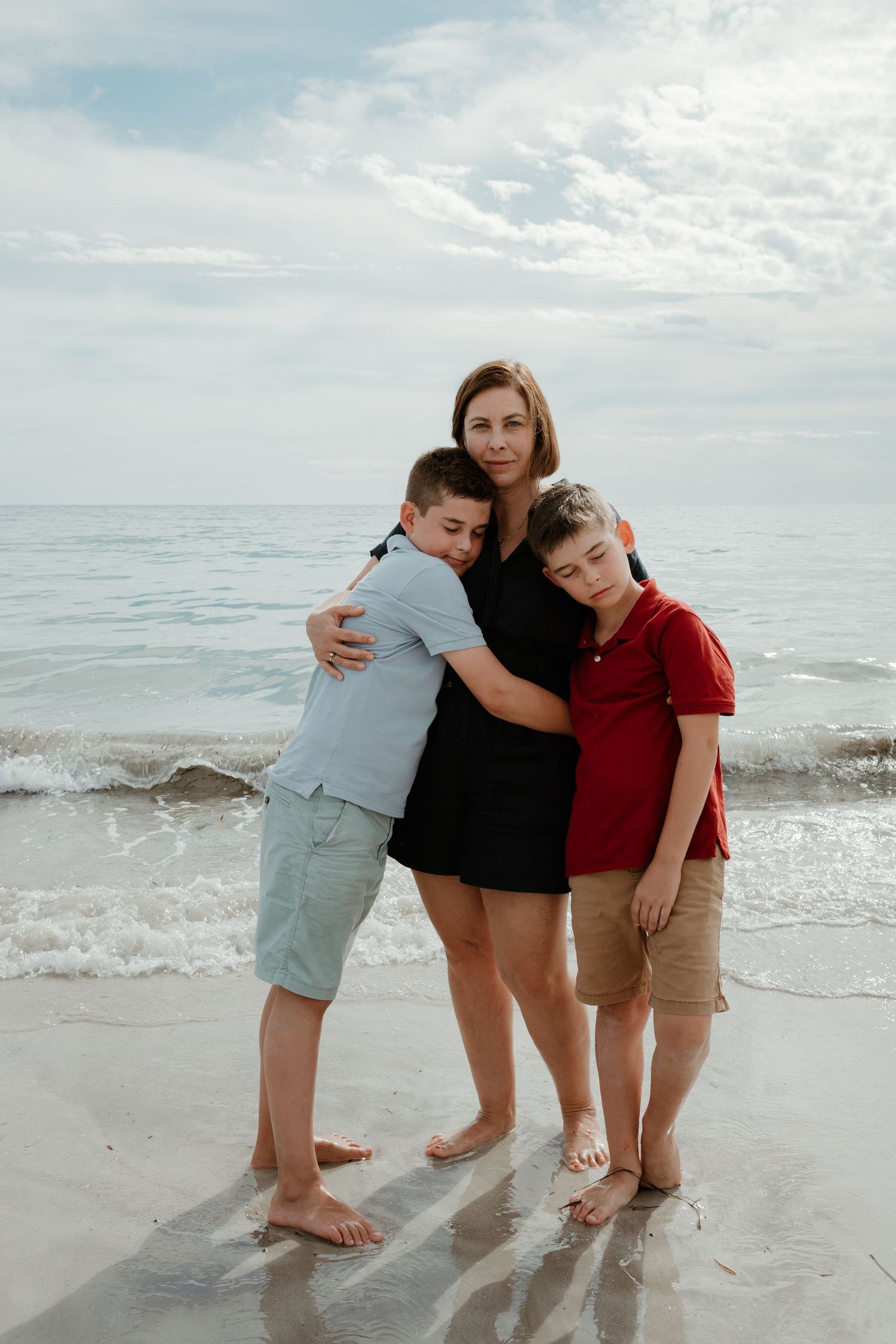 Three people embracing on a sunny beach with ocean waves in the background.