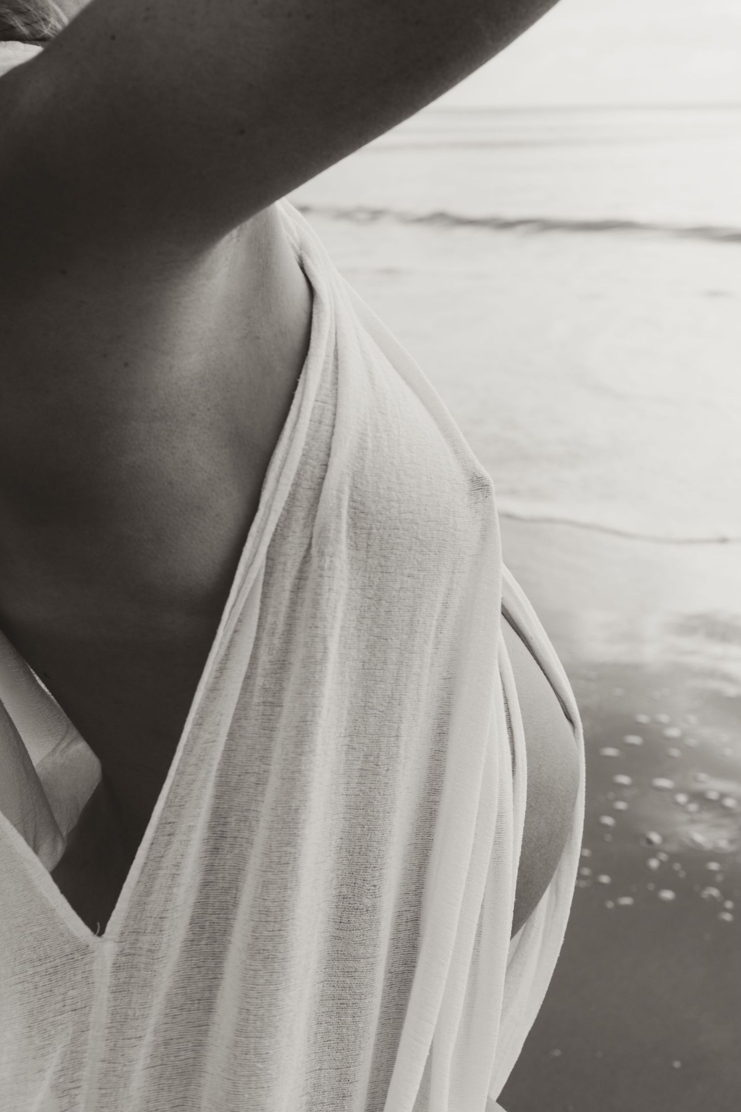 Black and white artistic photograph of a flowing white fabric draping over a shoulder against a beachy background.