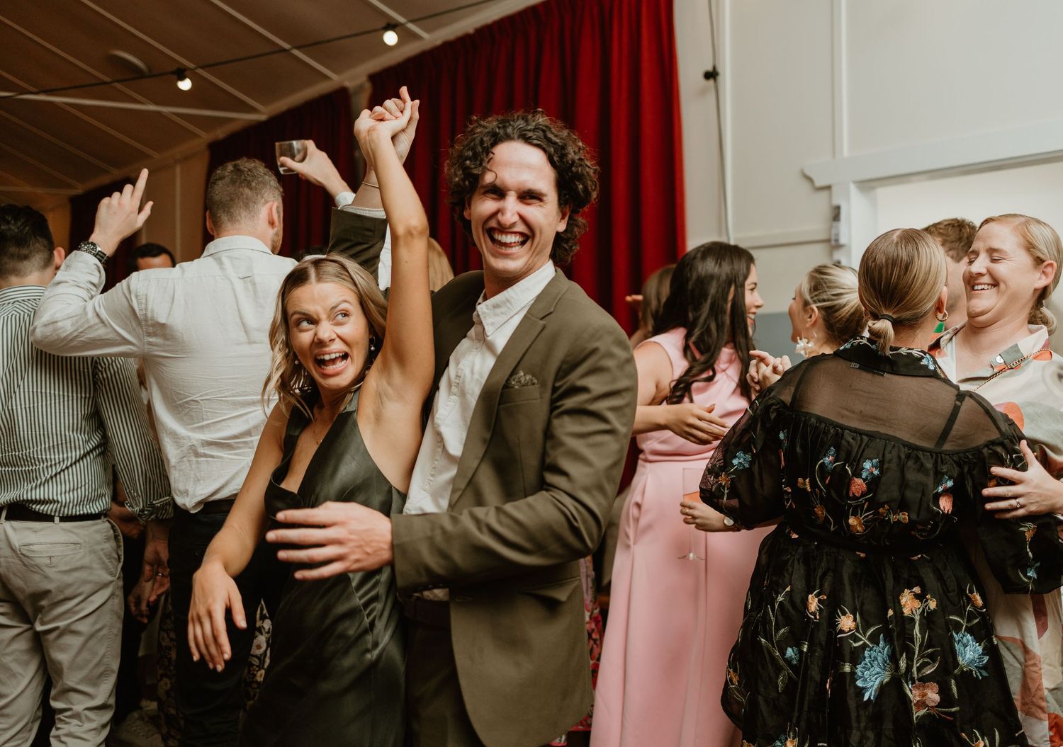 Wedding guests dancing joyously on a dance floor with red curtains in the background.