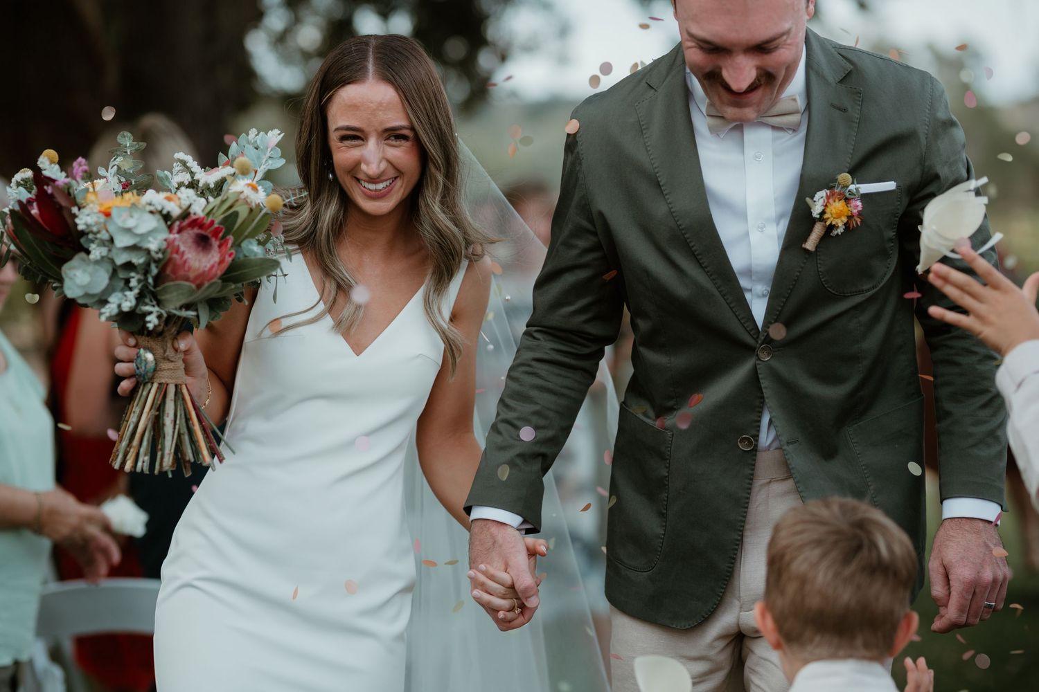 A couple walks hand in hand outdoors, with the bride in a white dress and groom in a green blazer.