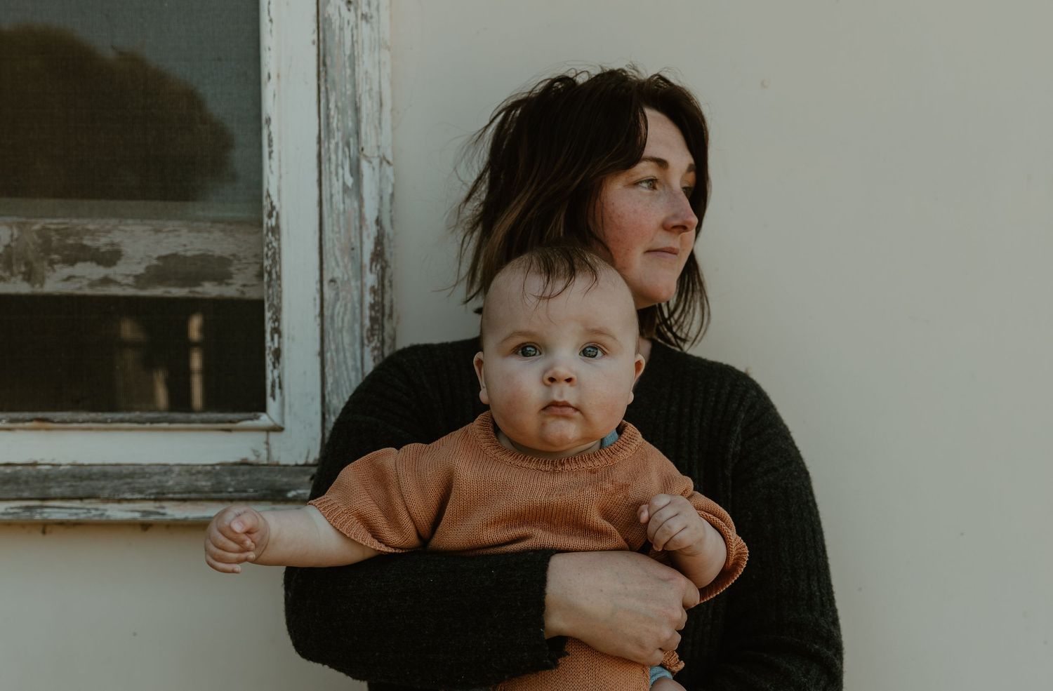 Parent holds baby wearing orange sweater while sitting next to weathered window frame.