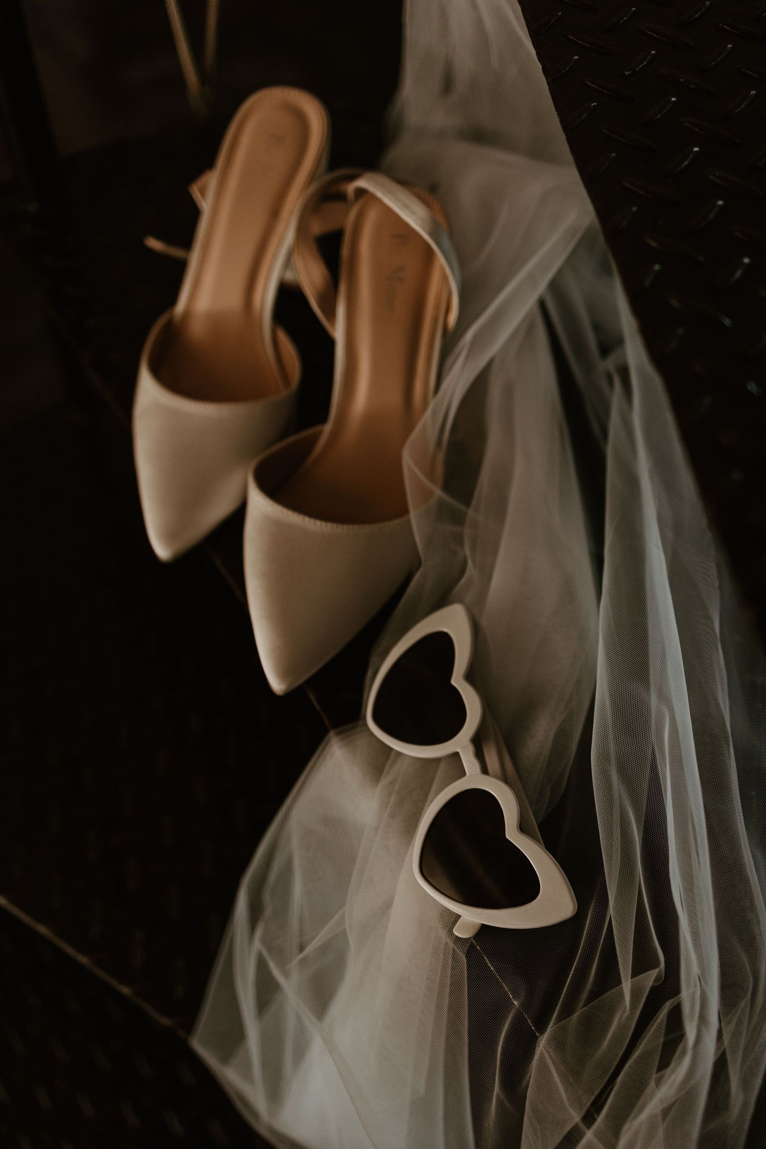 Wedding accessories including beige heels, heart-shaped sunglasses and delicate veil on dark surface.
