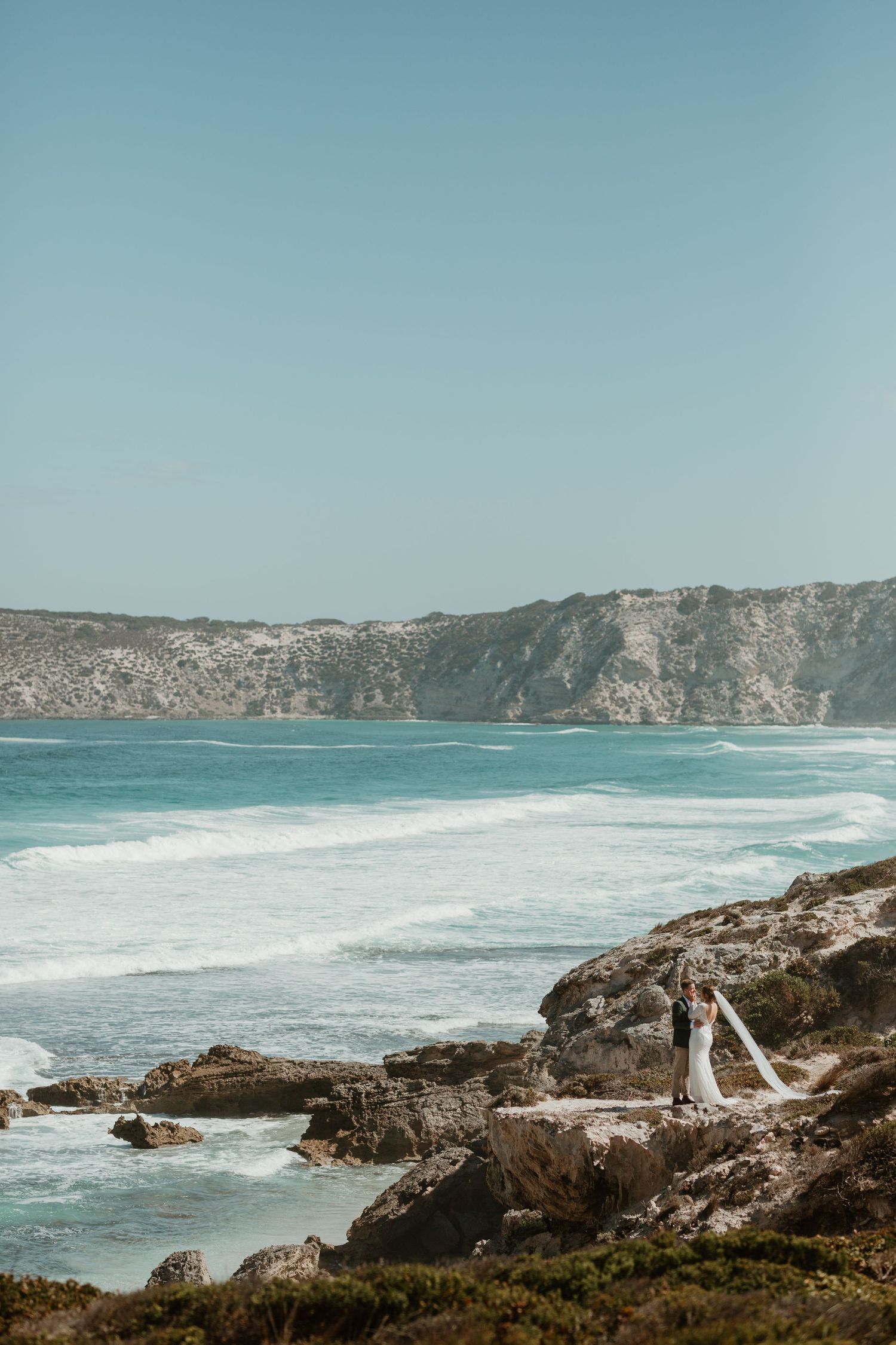 Couple in wedding attire standing on dramatic coastal cliffs overlooking turquoise ocean.