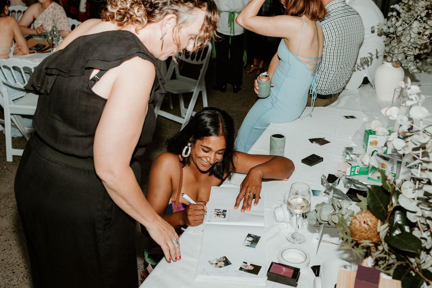 Guests signing a wedding guestbook at a decorated reception table with flowers and drinks.