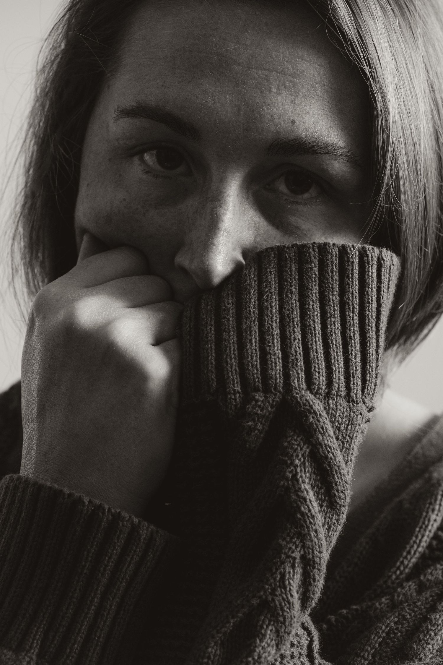 A dramatic black and white portrait showing someone partially hiding their face behind a ribbed knit sweater sleeve.