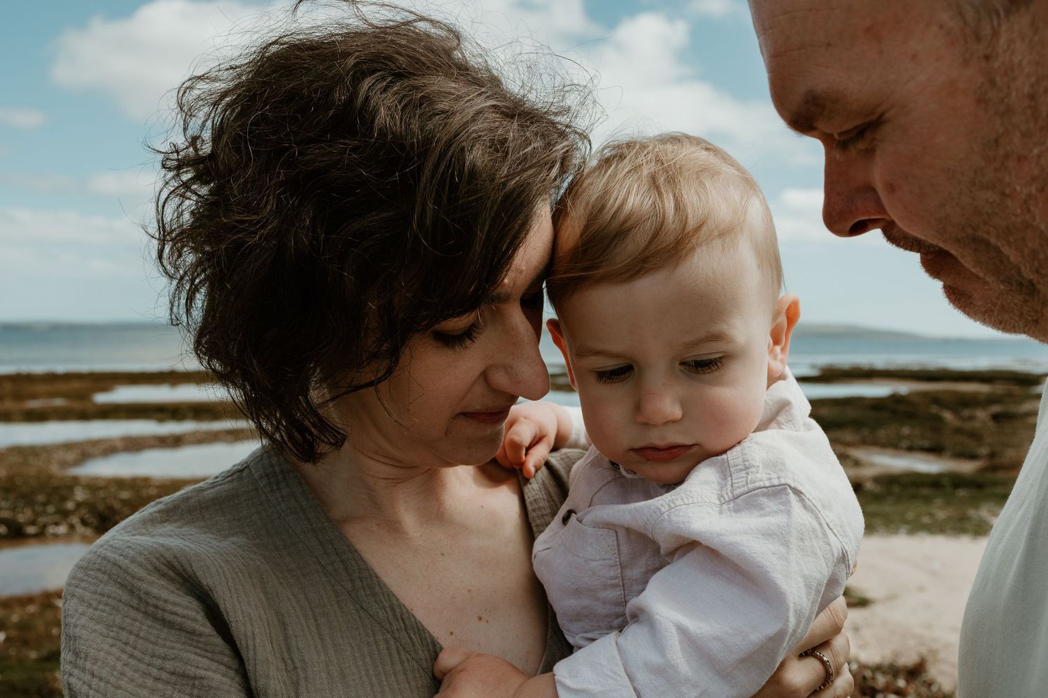 Close-up intimate portrait of parents with baby against coastal landscape.