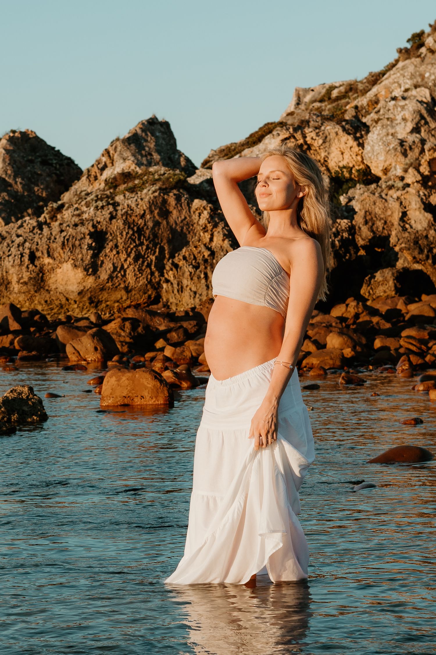 Figure standing in shallow ocean water at sunset against rocky cliffs wearing white flowing skirt and crop top.