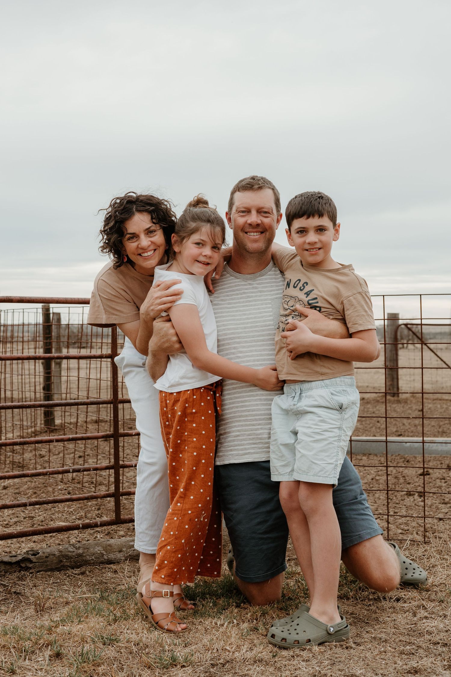 Family of four embraces while posing together against rural fence backdrop.
