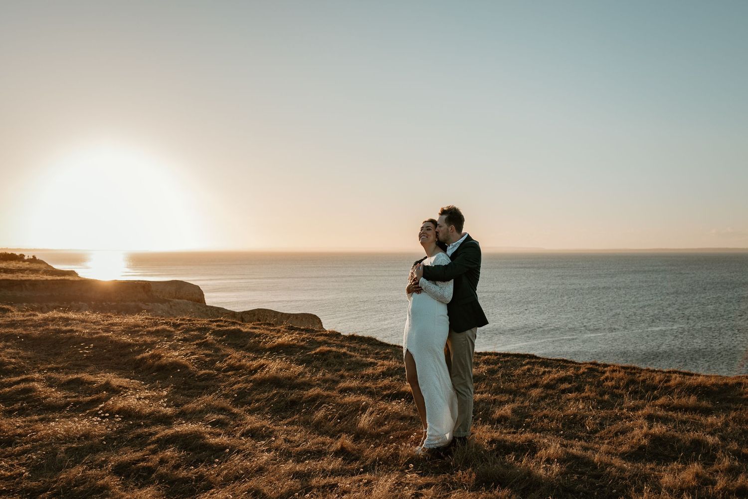 Couple embracing on coastal cliff edge during golden sunset over ocean.