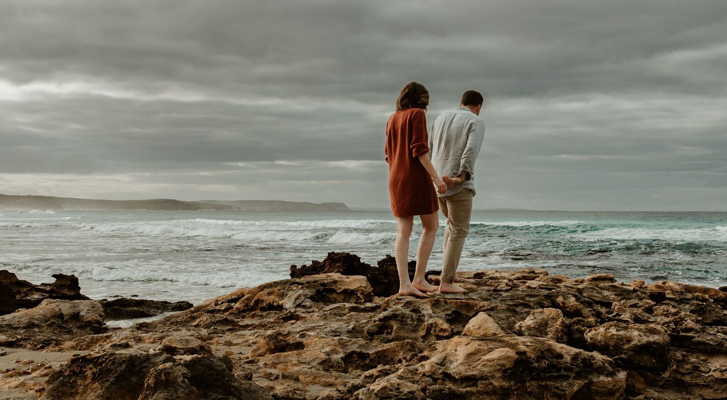 Two figures stand on rocky Kangaroo Island coastal outcrop overlooking moody ocean under cloudy sky at sunset.
