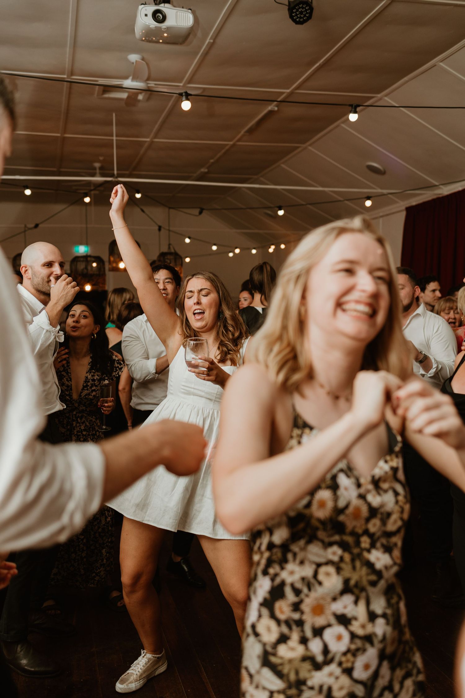 Wedding guests dancing energetically at an indoor reception with string lights overhead.