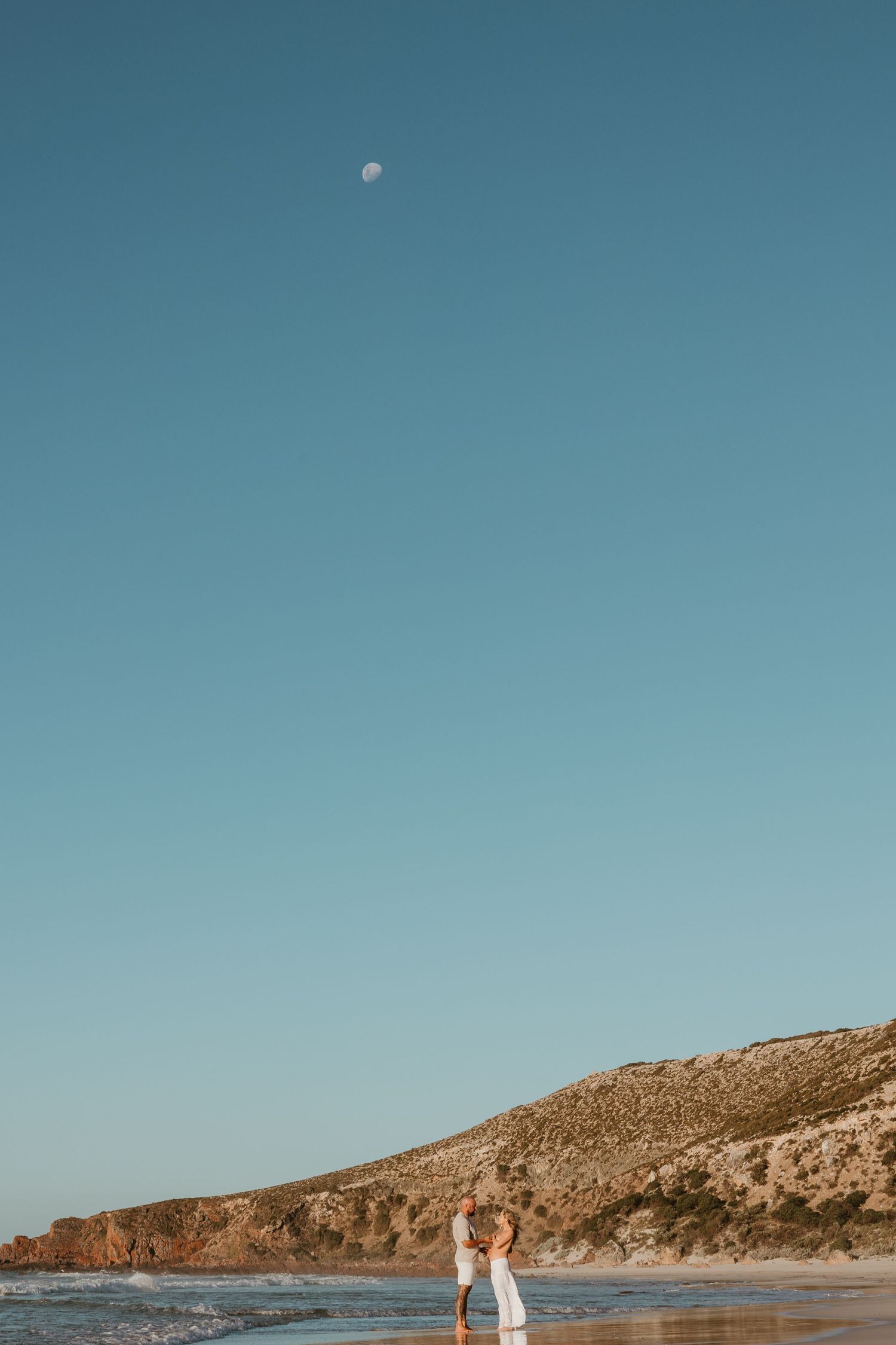 Moon visible in blue sky above coastal cliffs and beach landscape.