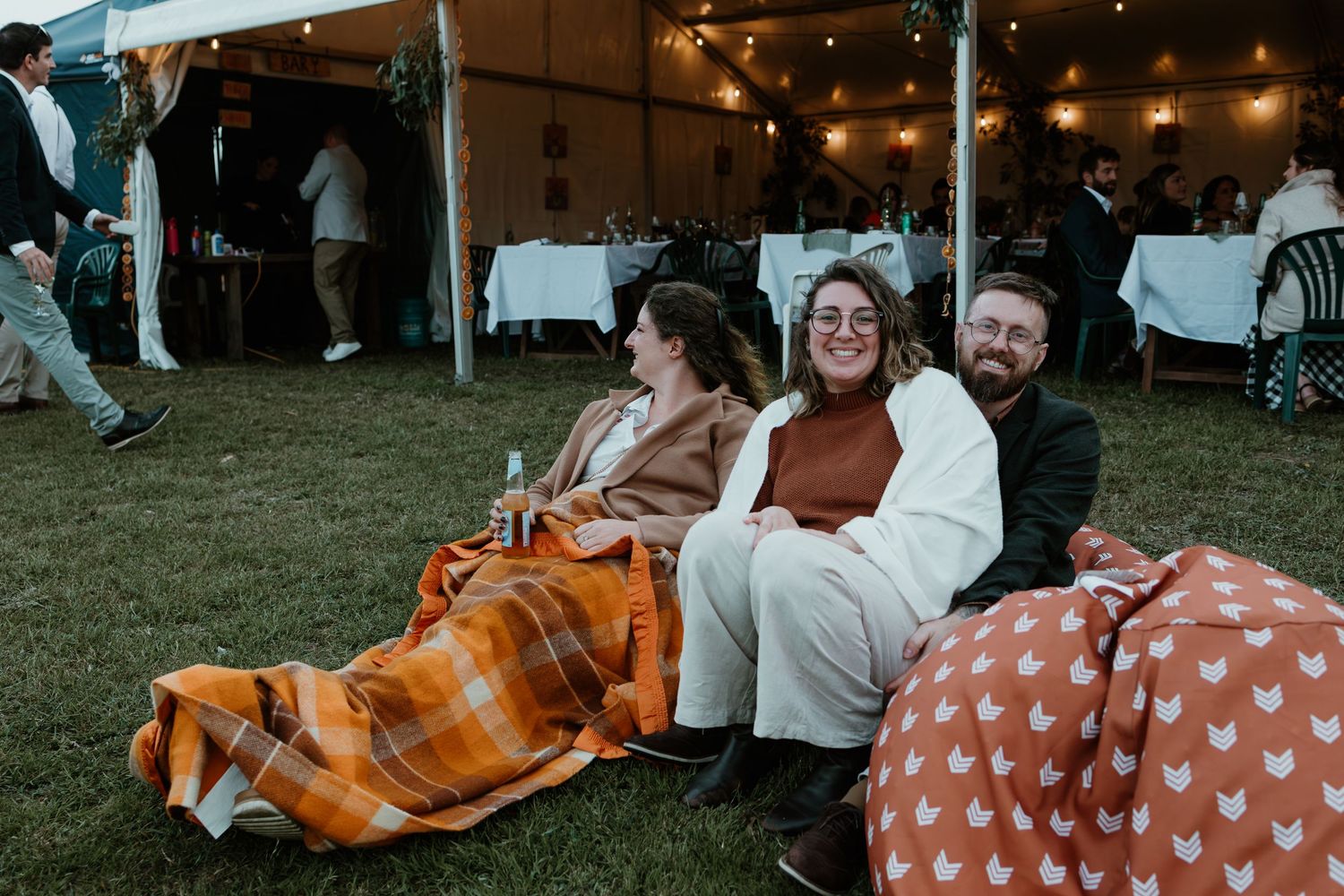 Wedding guests relax on blankets under a tent during an outdoor evening celebration.