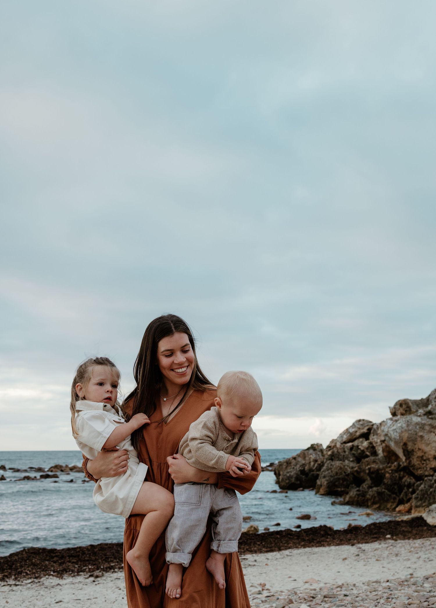 Woman holding two small children on rocky beach during cloudy sunset.
