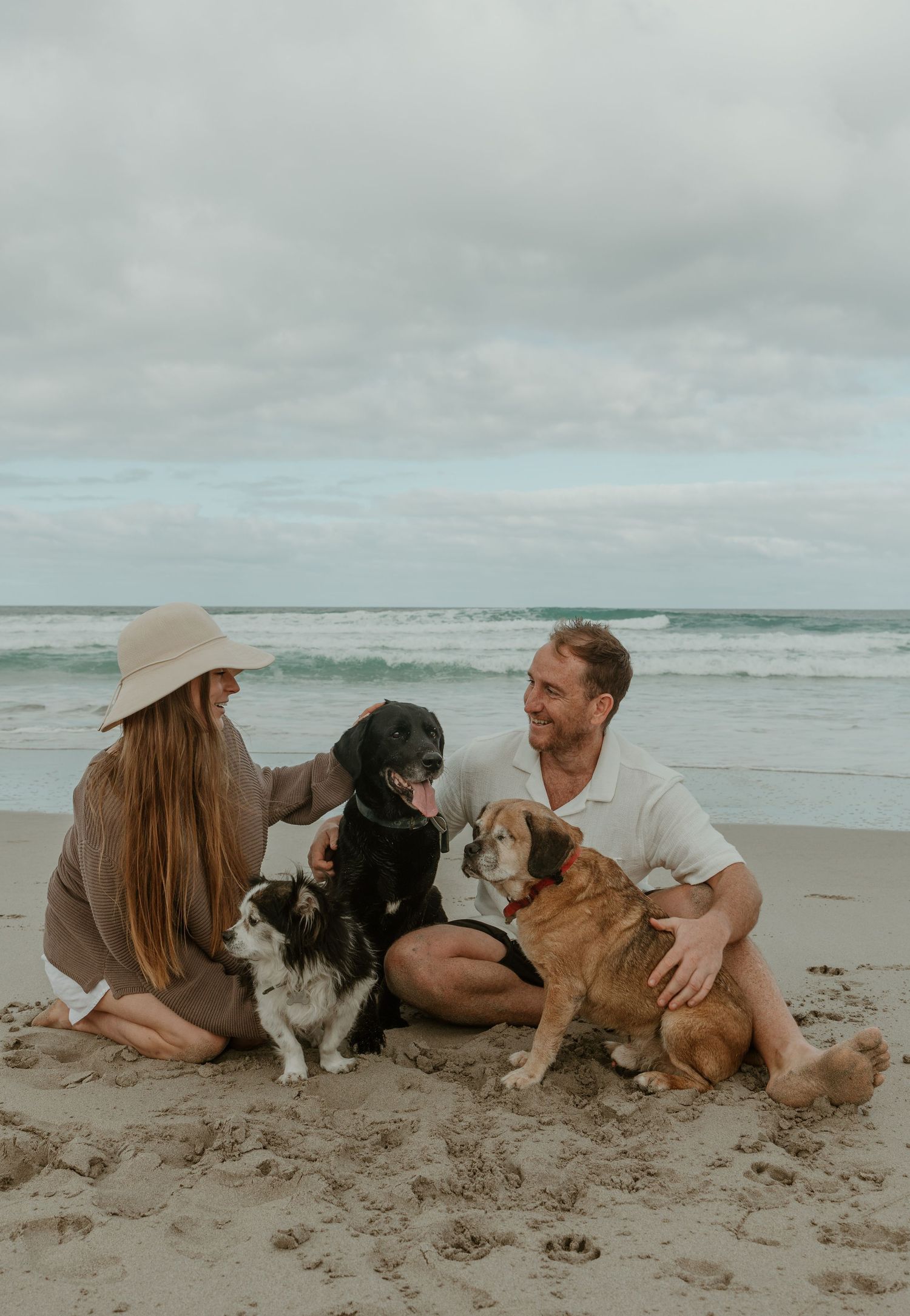 Couple sits on beach with three dogs while enjoying overcast coastal scenery.