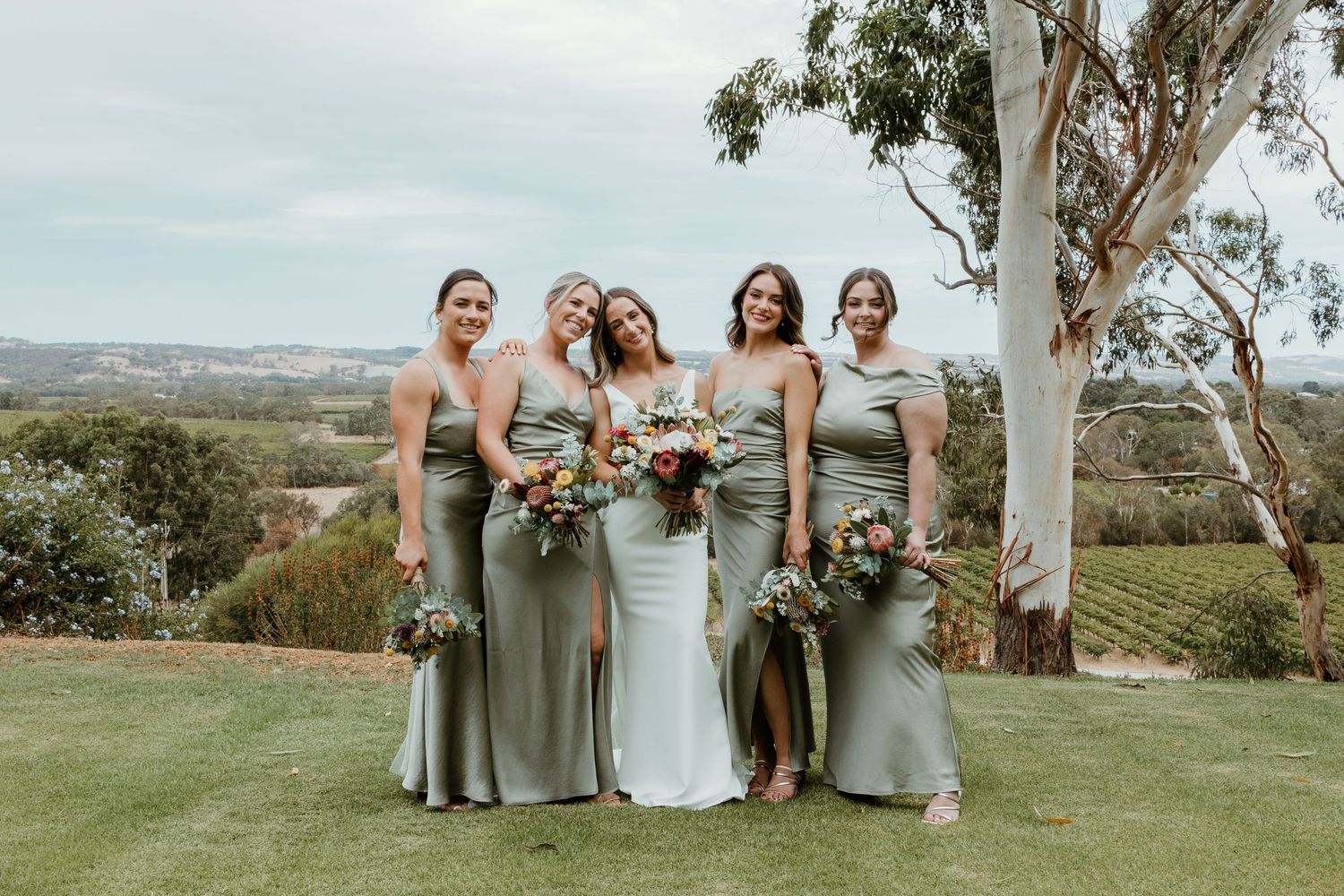 Bridesmaids in sage green dresses pose with bouquets on a scenic hilltop.