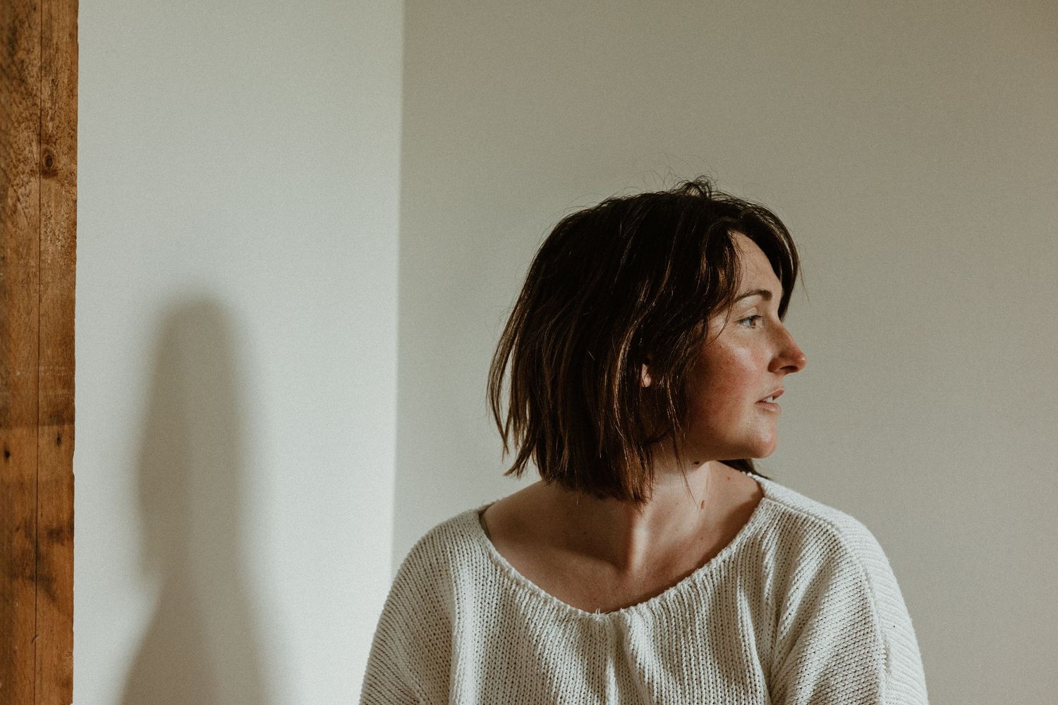 Person in white knit sweater with short dark hair looking away contemplatively against a white wall.