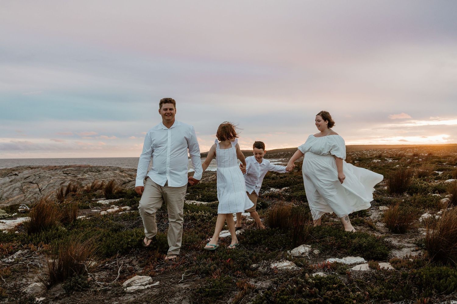 Family of four walking along rocky coastal terrain at sunset with flowing white dresses in the breeze.