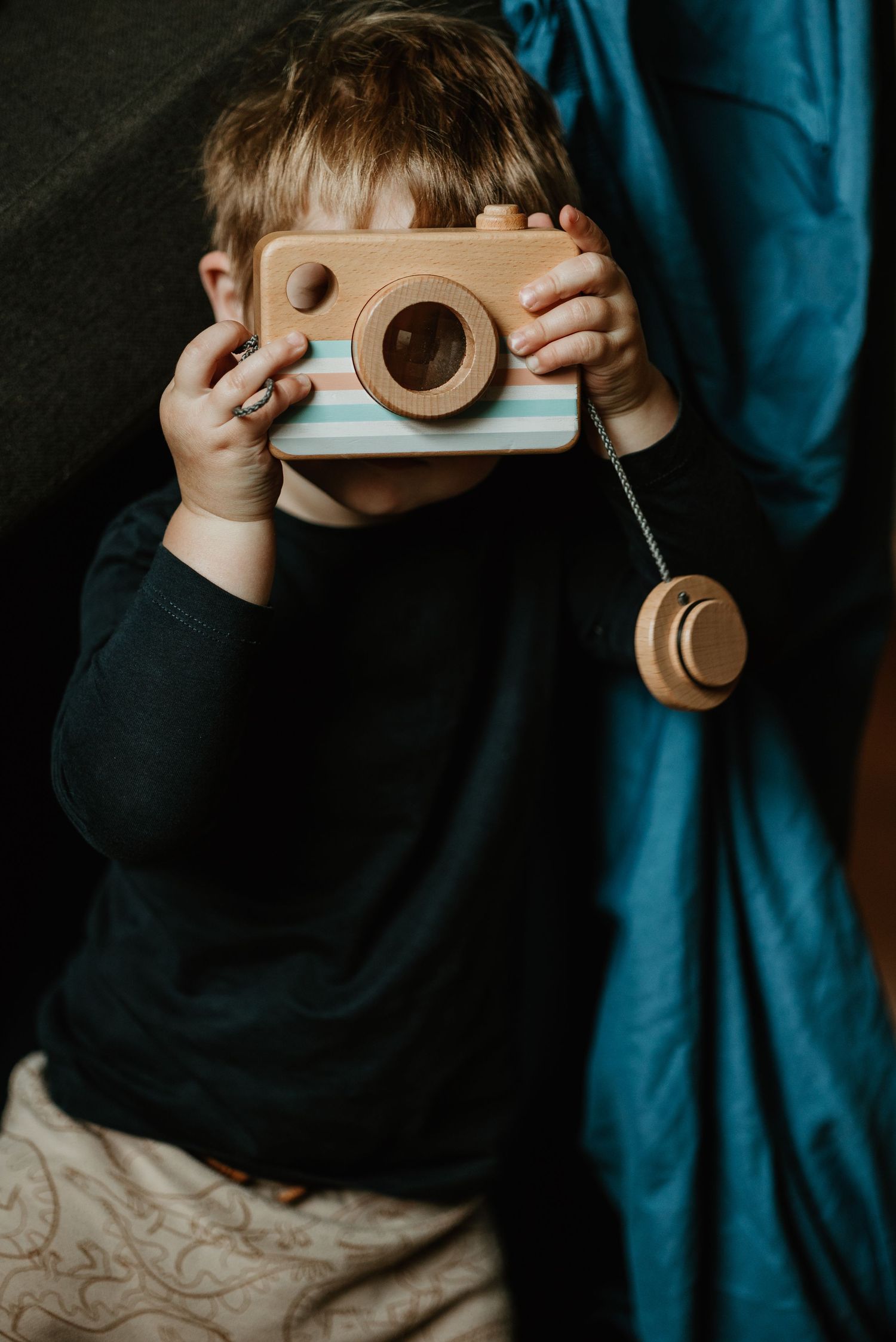 Child holding wooden toy camera against dark background with blue fabric. Kangaroo Island.