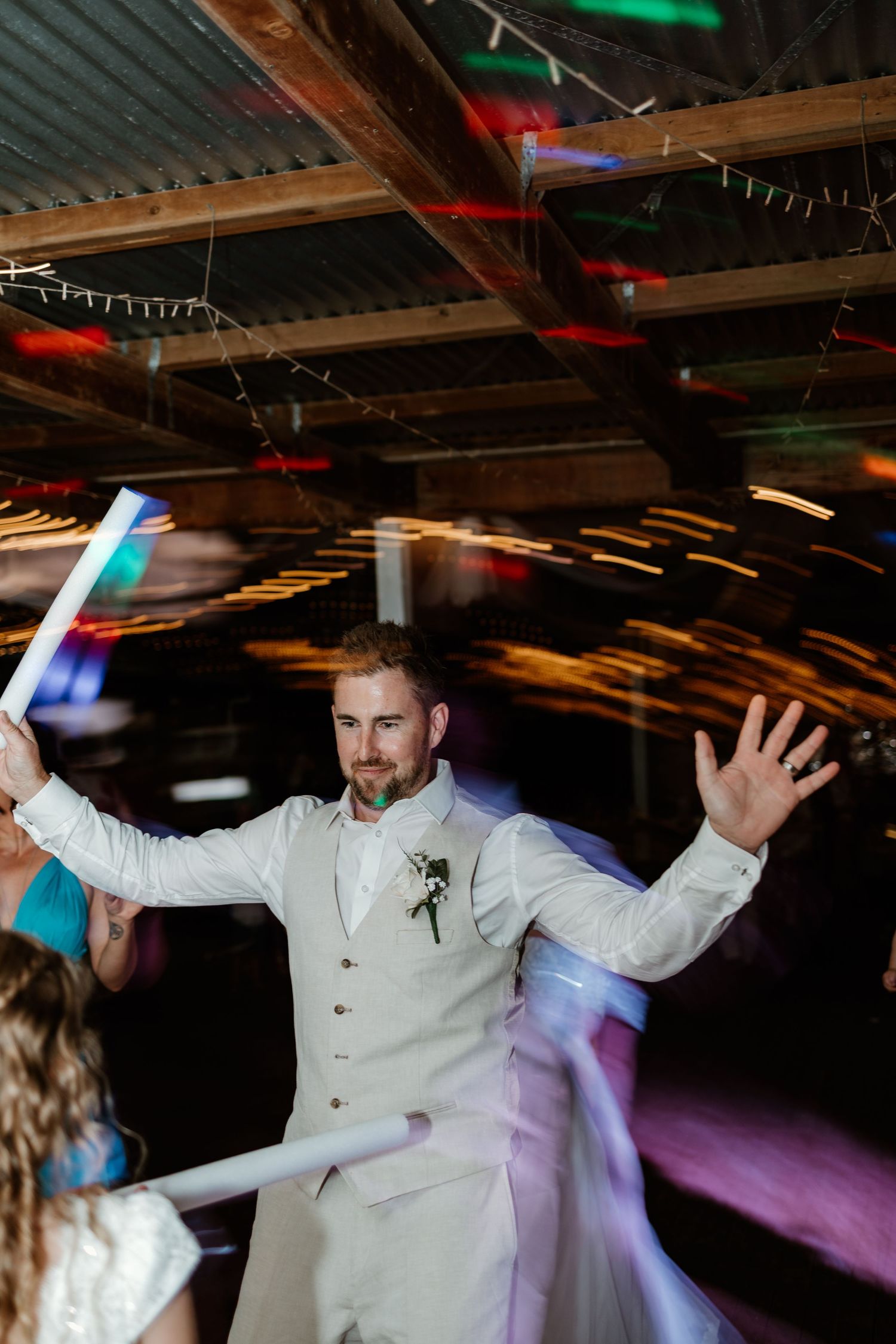 Groom dancing enthusiastically in a white suit under colorful party lights at wedding reception.