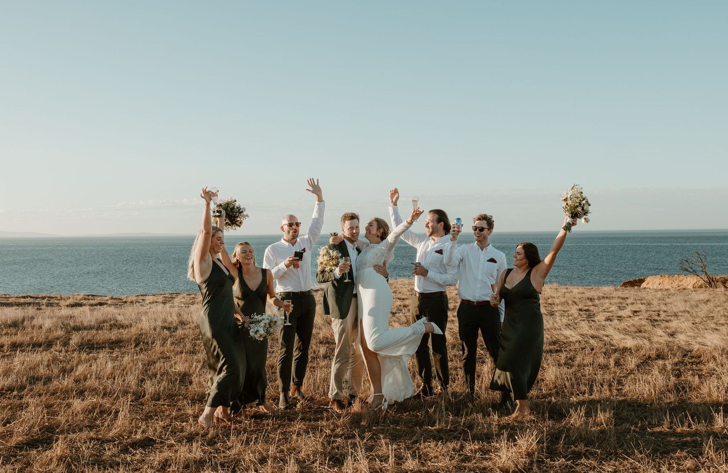 Wedding party celebrates joyfully on a coastal cliff overlooking the ocean.