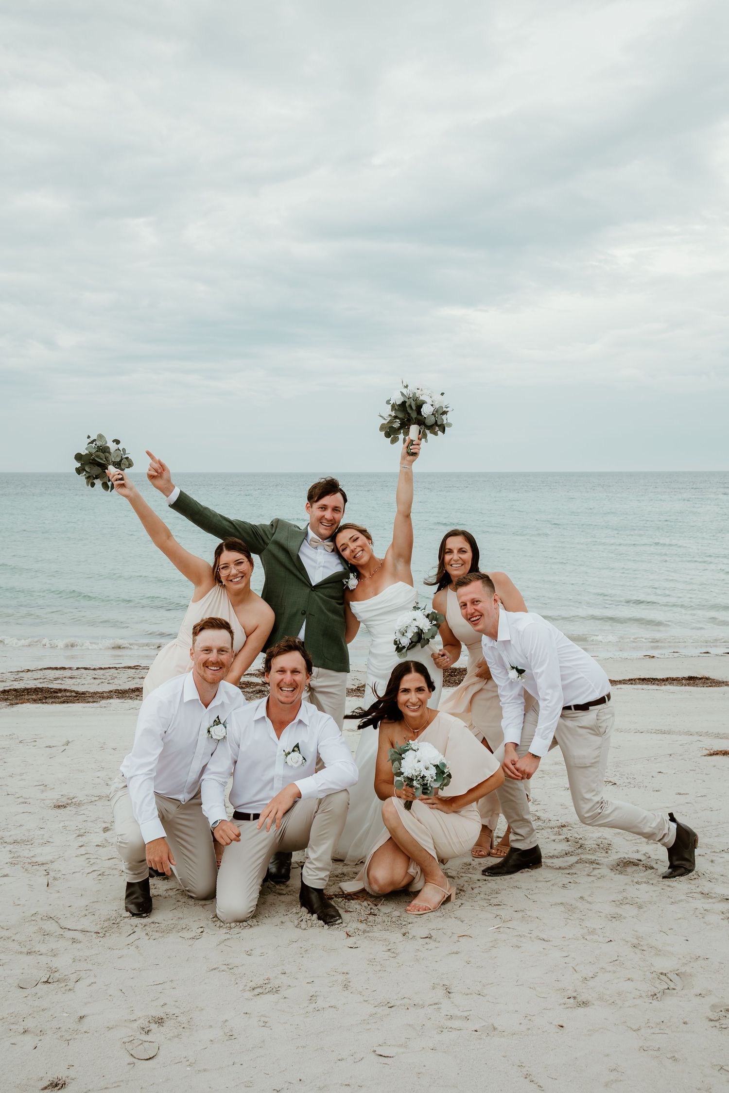 Wedding party celebrating on a beach, bride and groom lifted by groomsmen and bridesmaids holding bouquets.