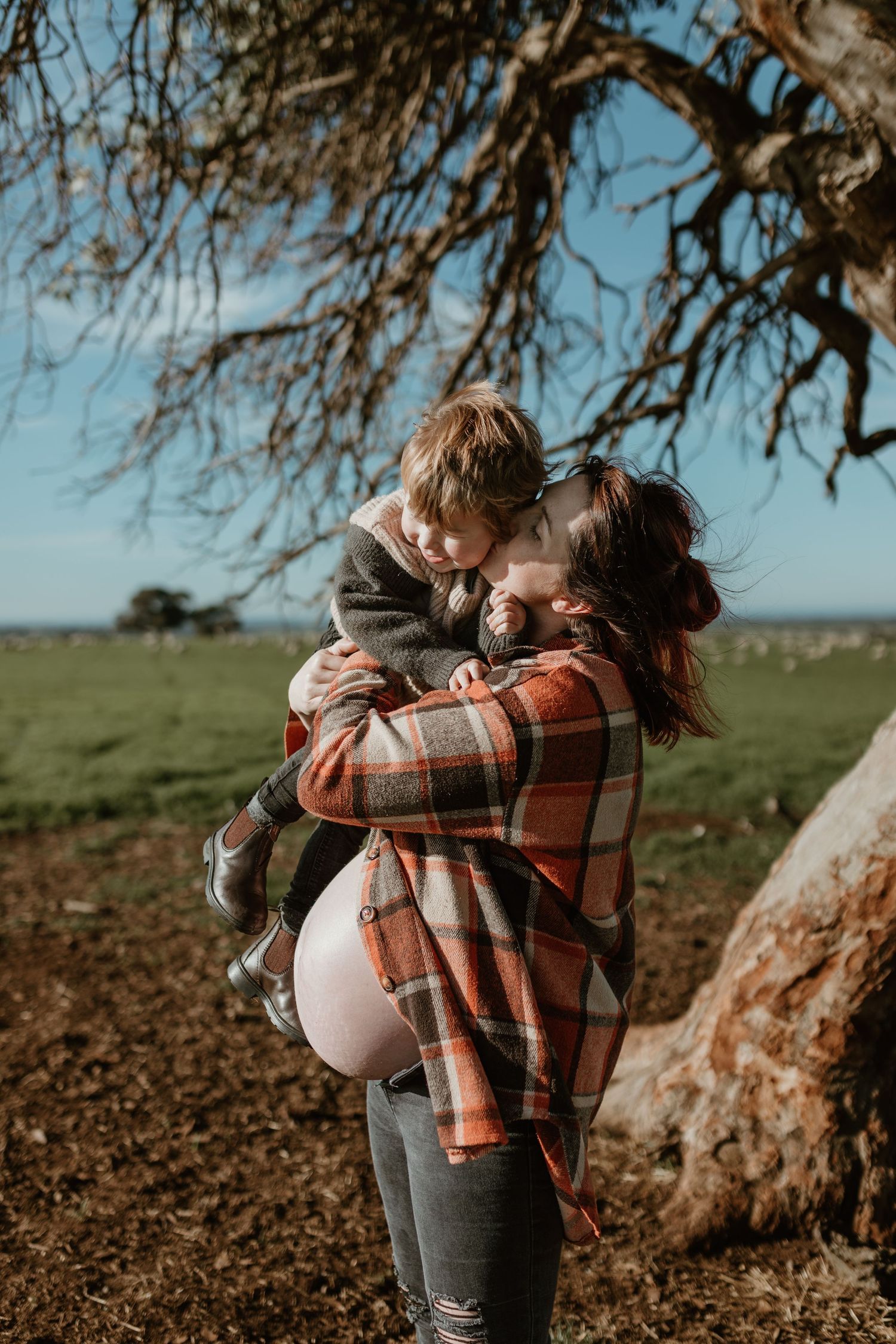 A tender outdoor moment captured against a rustic tree backdrop and green field on a sunny afternoon.