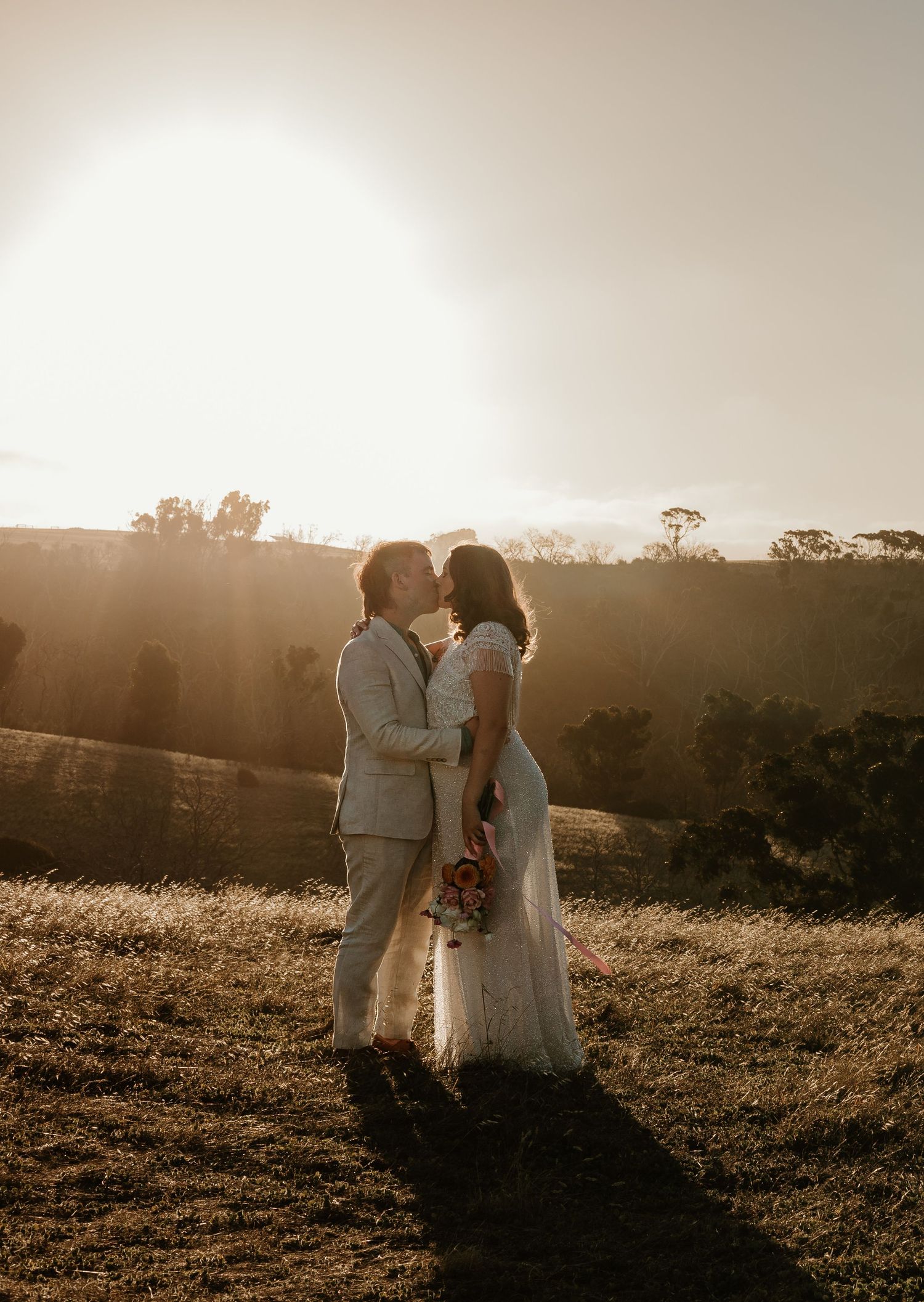 Newlyweds embrace in golden sunset light on a hilltop meadow.