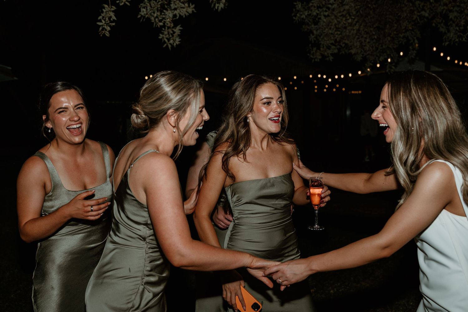 Bridesmaids in sage green dresses laugh and dance together at an evening wedding reception.