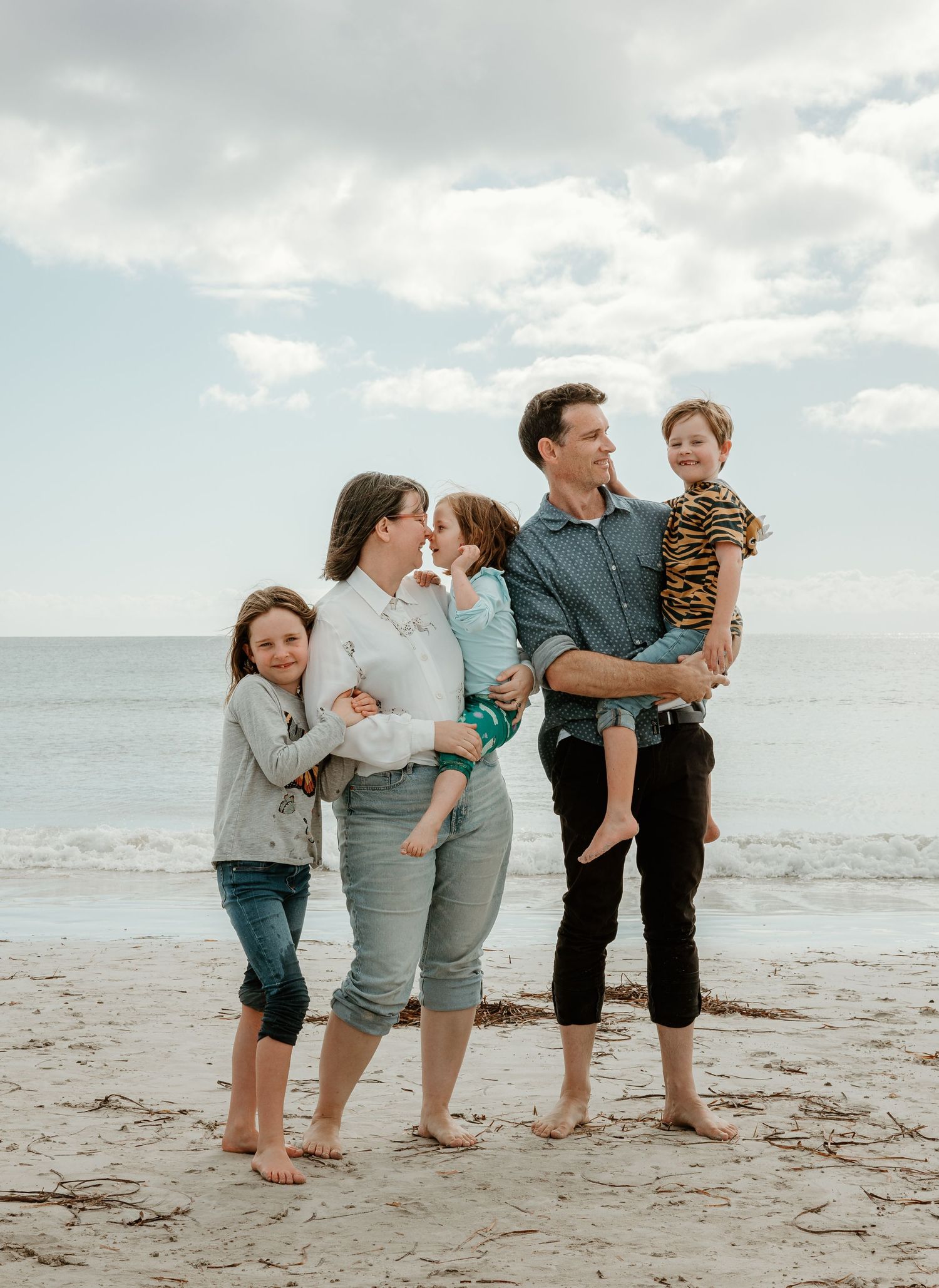 Barefoot family portrait on sandy beach with ocean and cloudy sky background.