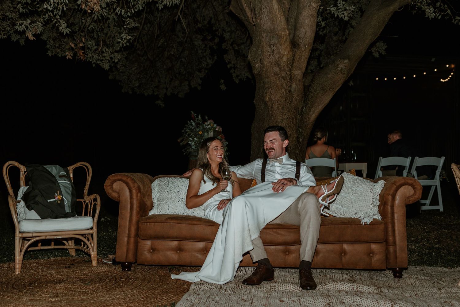 Couple sits together on vintage leather couch under string lights at outdoor evening event.