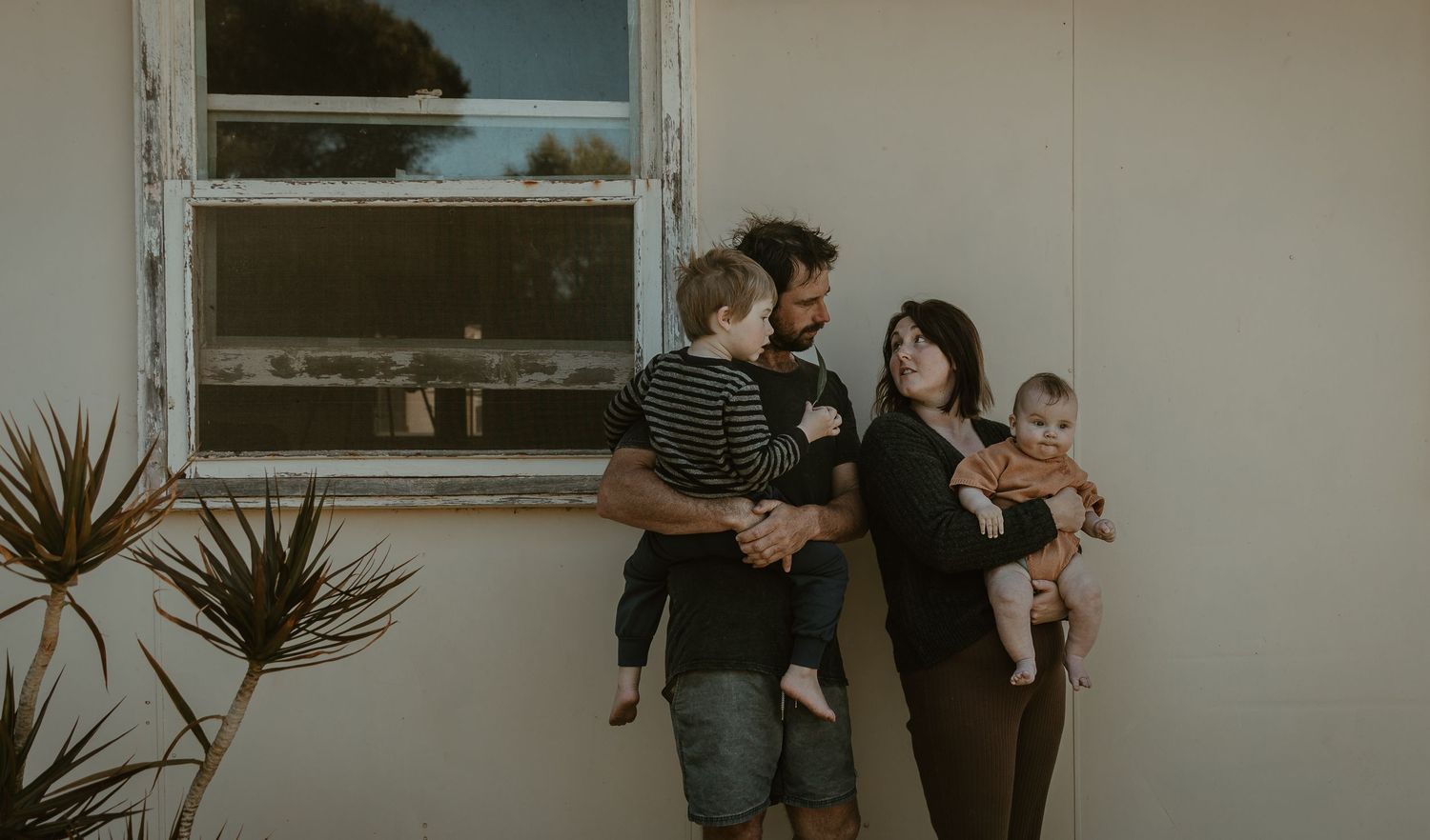 A couple stands with their babies against a wall with a window, surrounded by potted plants in warm lighting.