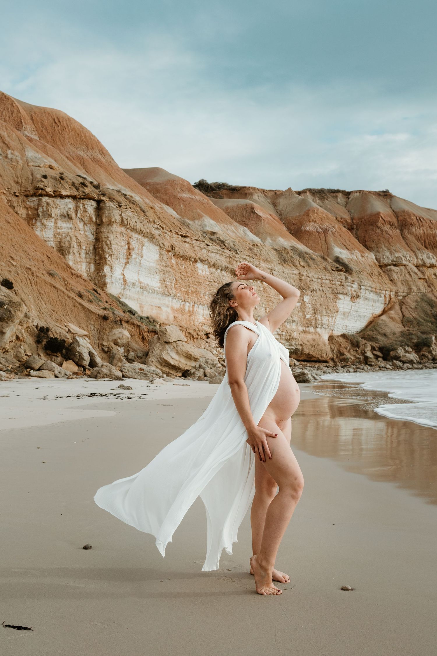 A flowing white dress billows in the coastal breeze as someone poses on a sandy beach with dramatic cliffs in the background.