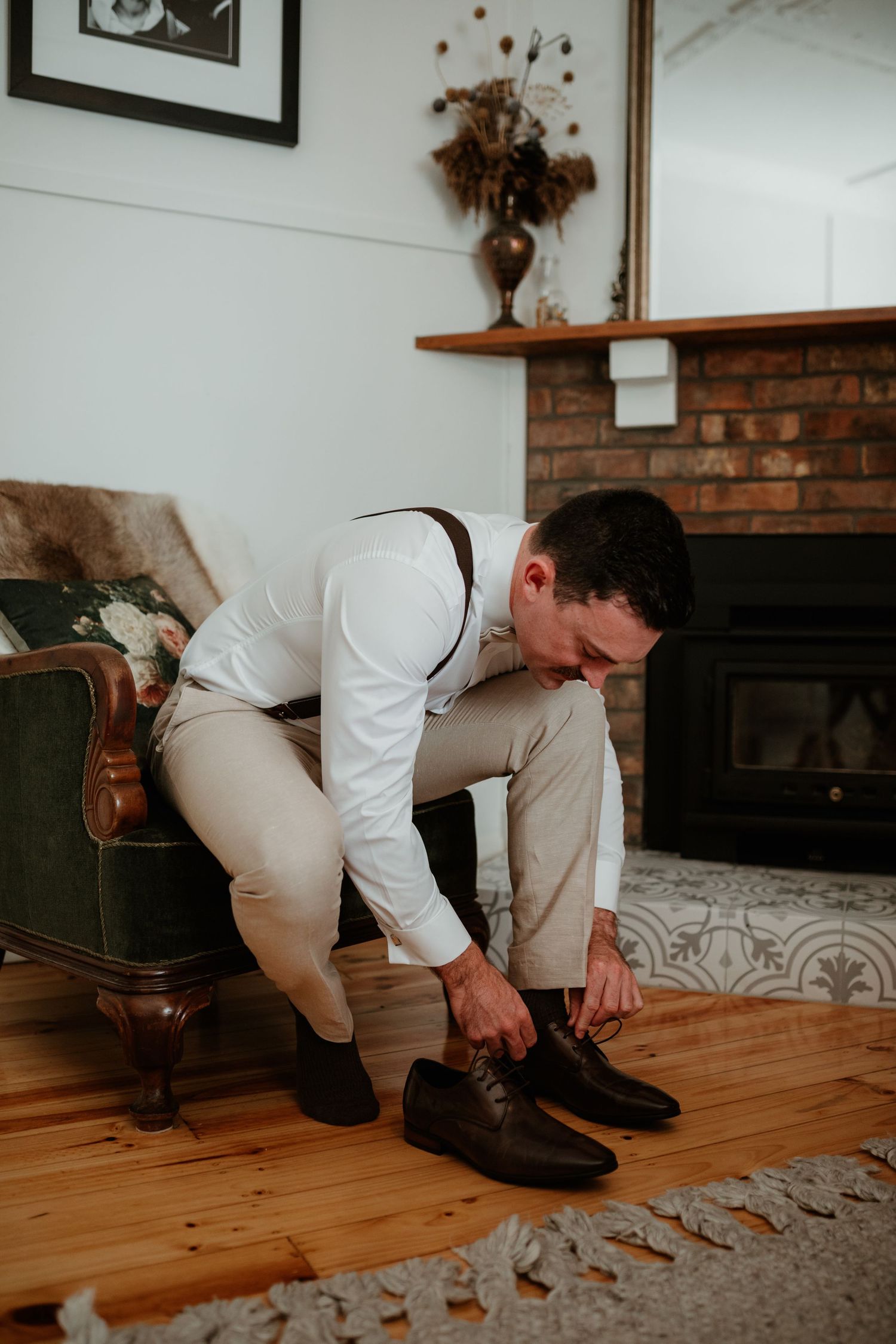 A person in formal attire ties their shoes while seated near a brick fireplace.