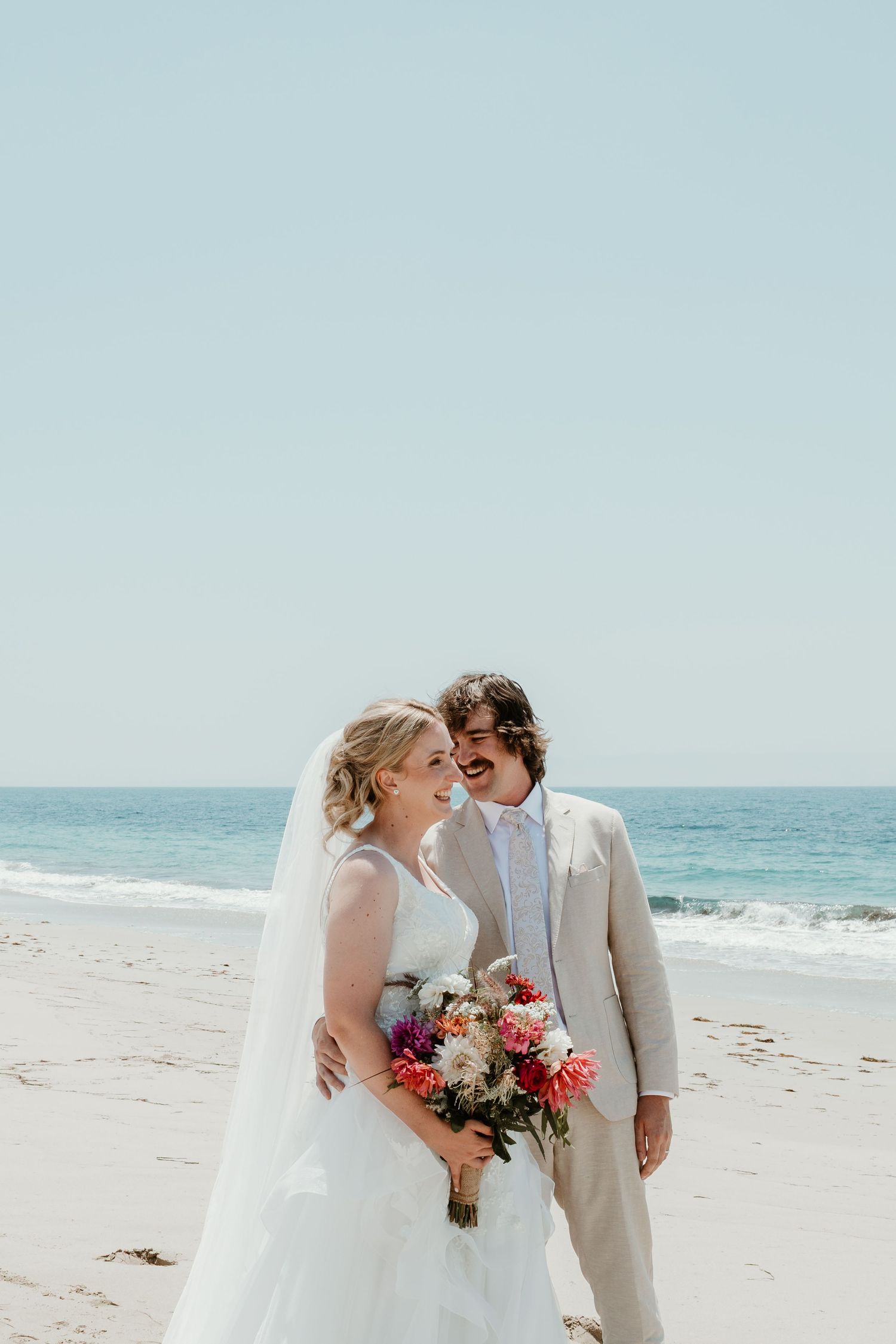 Newlyweds share joyful moment on sunny beach with bride holding vibrant bouquet.