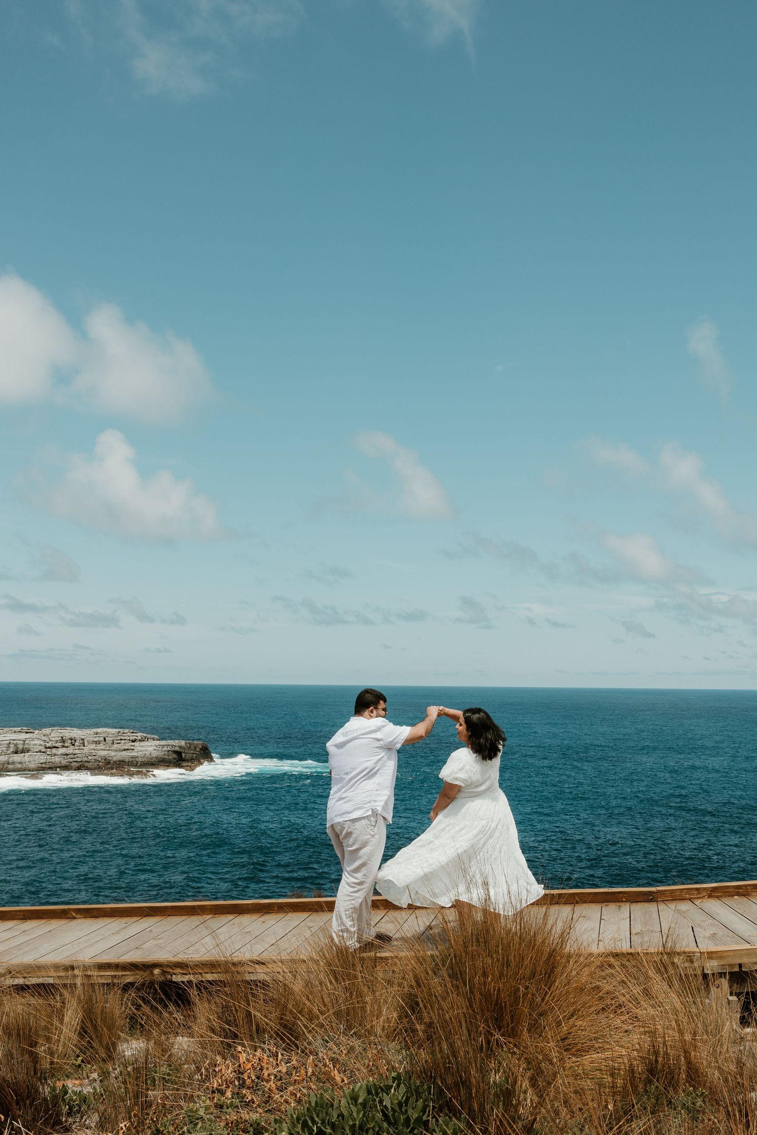 Couple dancing on wooden platform overlooking blue ocean waters.