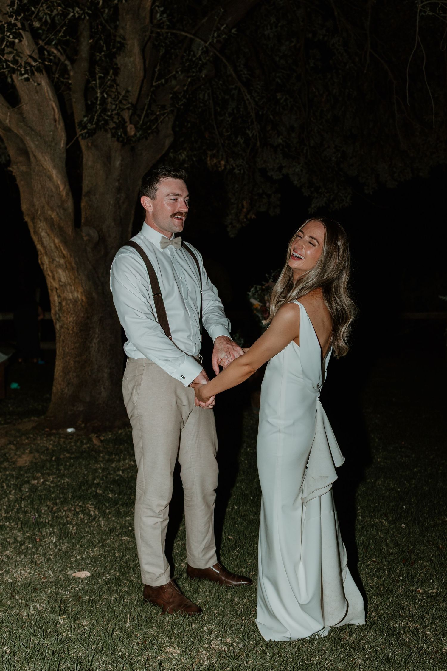A couple in light-colored wedding attire shares a joyful moment together in an outdoor garden setting at night.