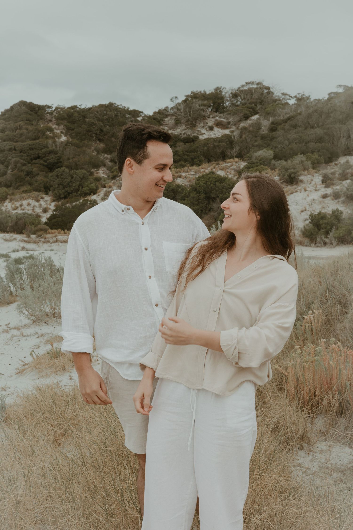 Couple in white clothing share tender moment on hilltop with coastal scenery.