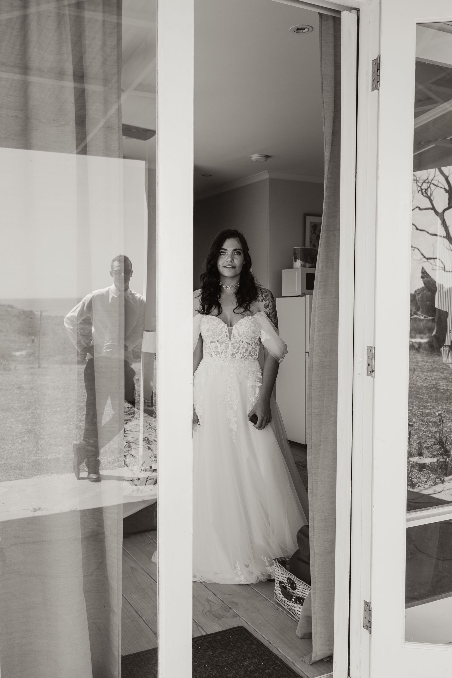 Black and white photo of a bride standing in a doorway wearing a flowing wedding gown.