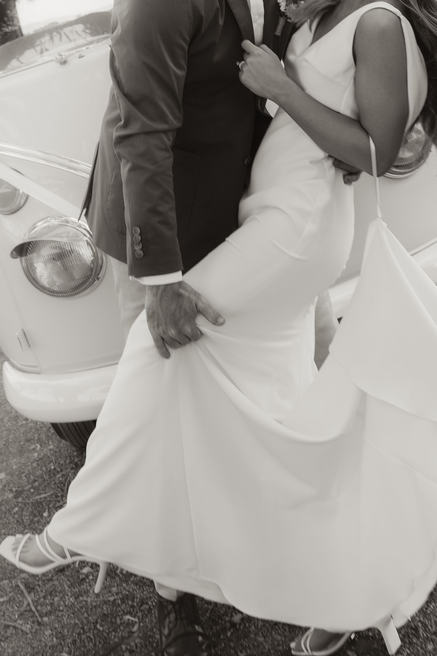 Black and white photo of couple in formal wear posing beside vintage car.