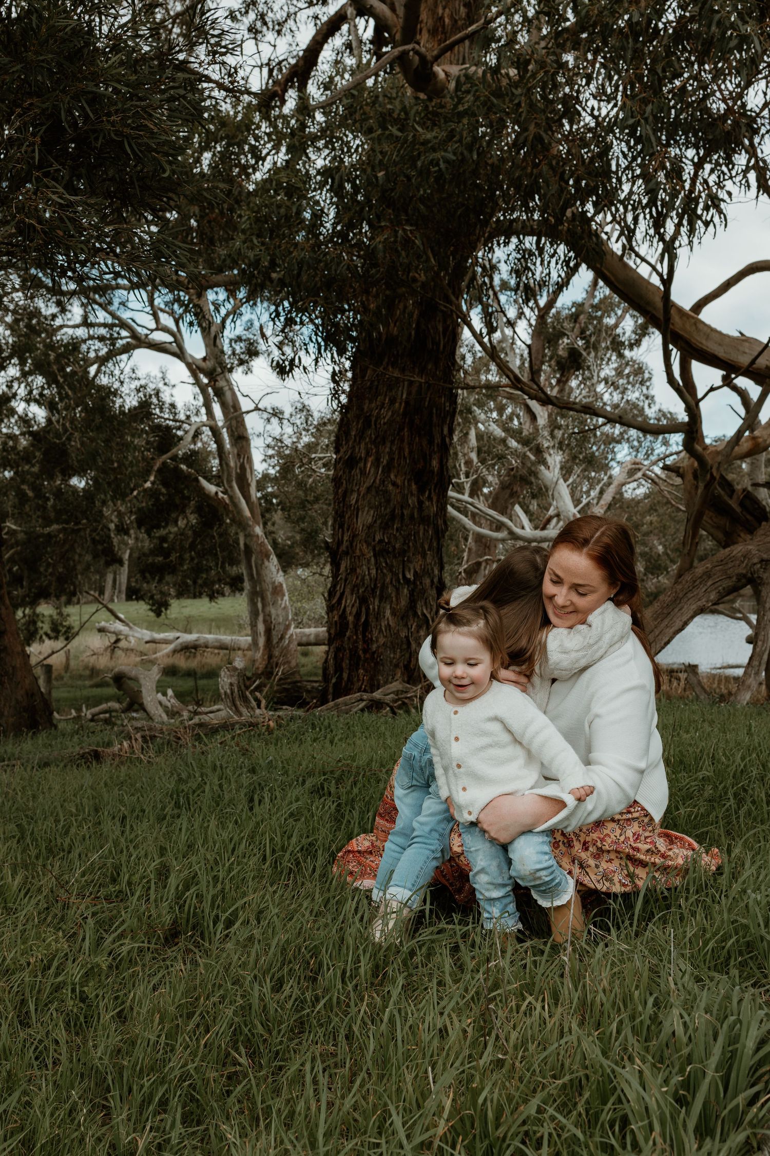A tender outdoor moment shared between Mother and daughters sitting in grass beneath tall eucalyptus trees.