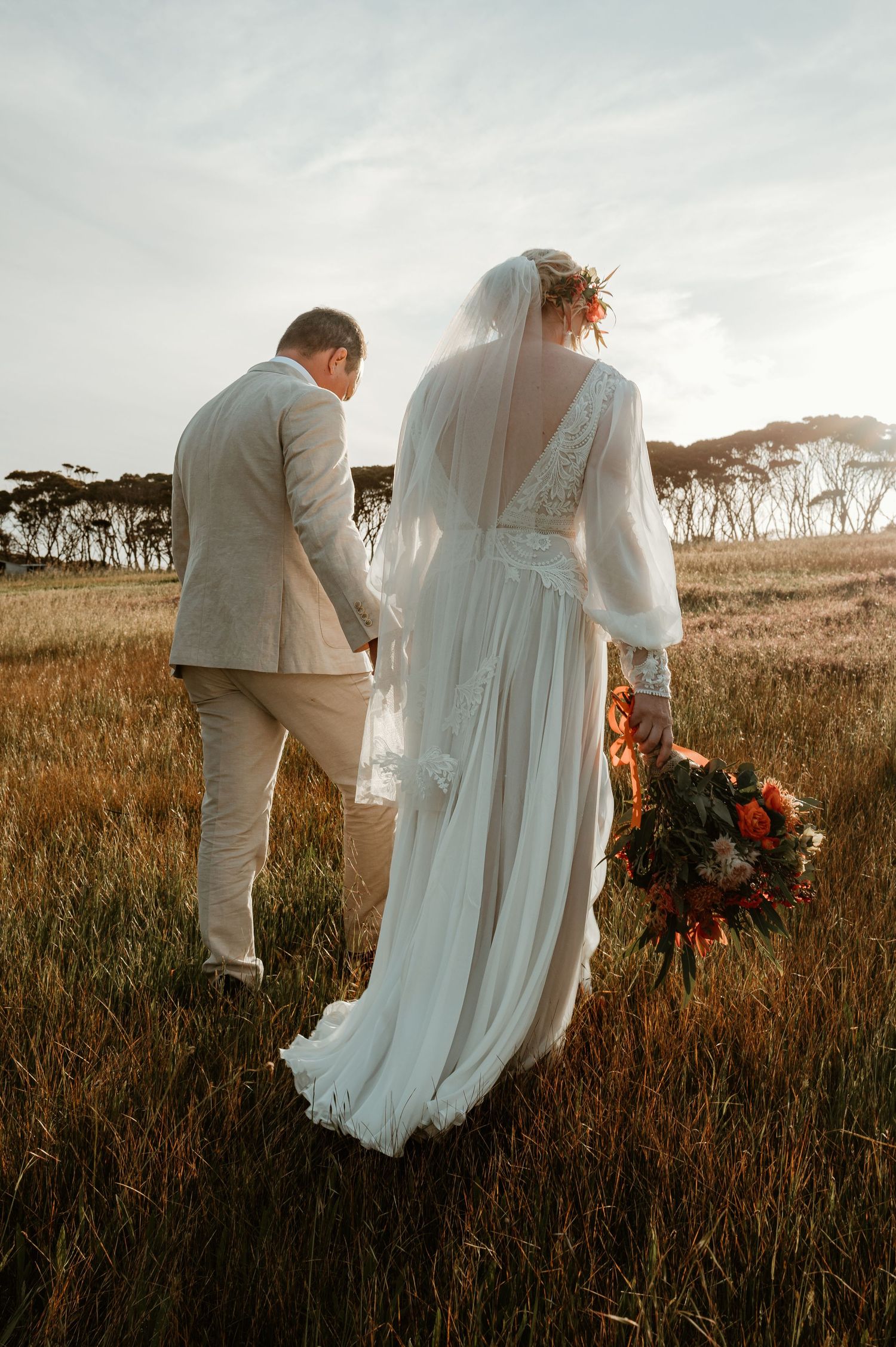 Bride in flowing white dress and groom walking through golden field at sunset.
