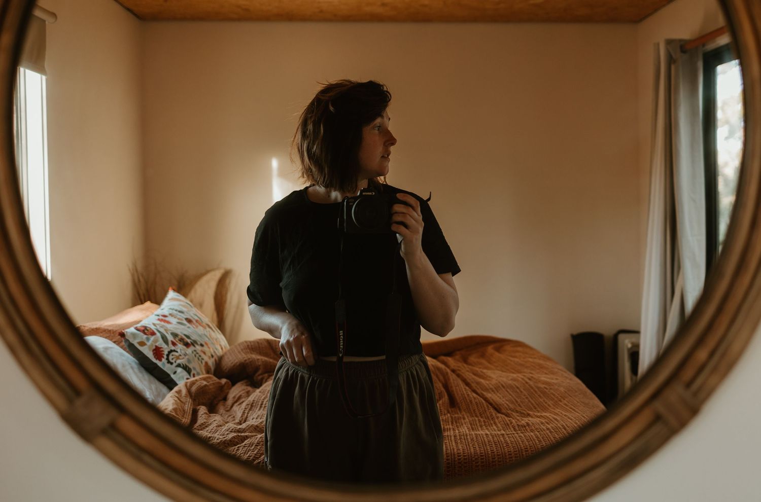 Mirror selfie in a warm-toned bedroom showing a figure in black dress taking photo with vintage oval frame.