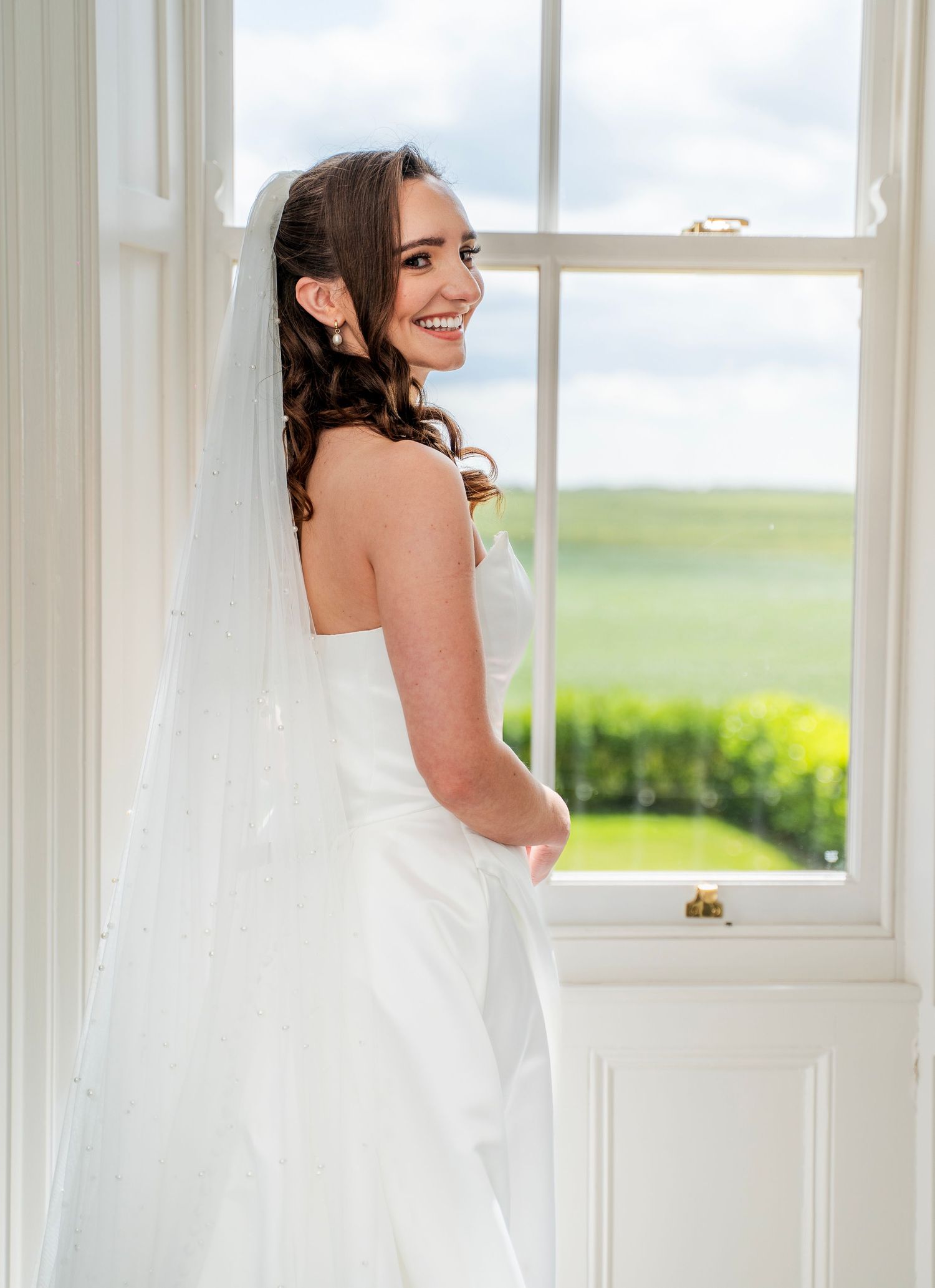 Bride in strapless white wedding dress with pearl-detailed veil smiling by a window at Carlton Manor, captured in candid wedding photography